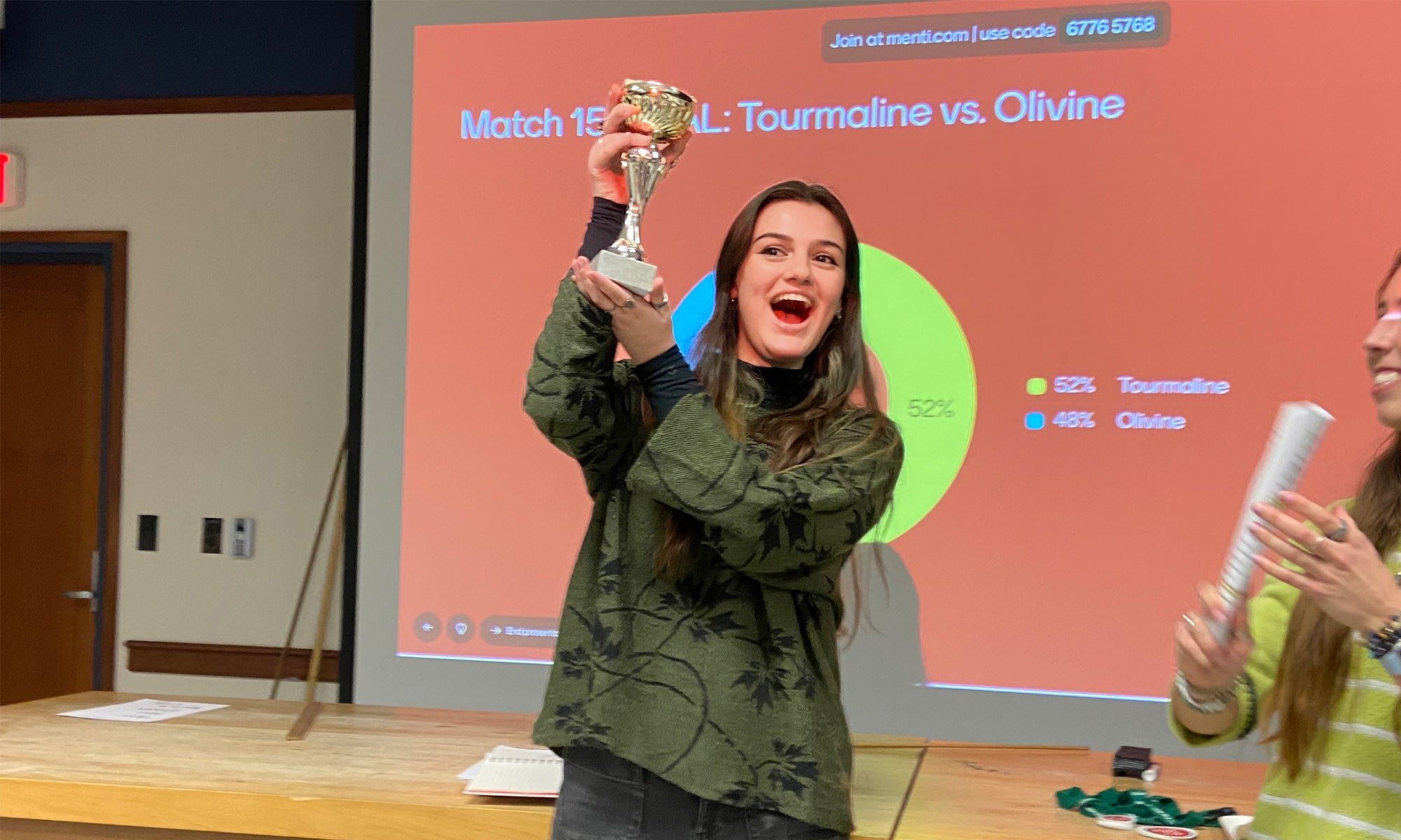 A student standing in front of a presentation projected on a screen holds up a trophy in celebration.