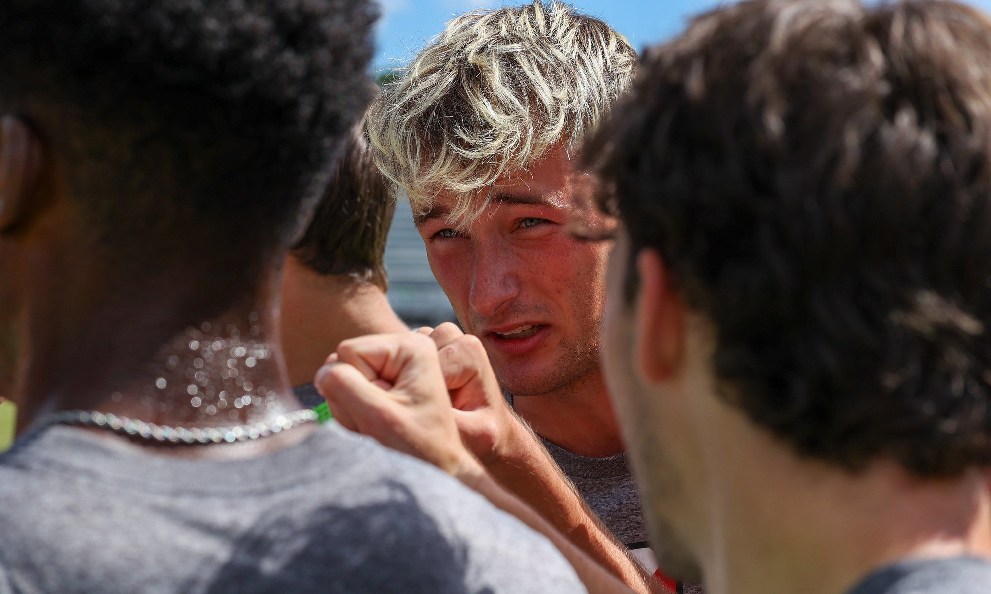 A close up photo of a group of college soccer players on the field; the face of a young man with blond hair is in focus in the center of the photo, surrounded by his teammates giving a celebratory fist bump.