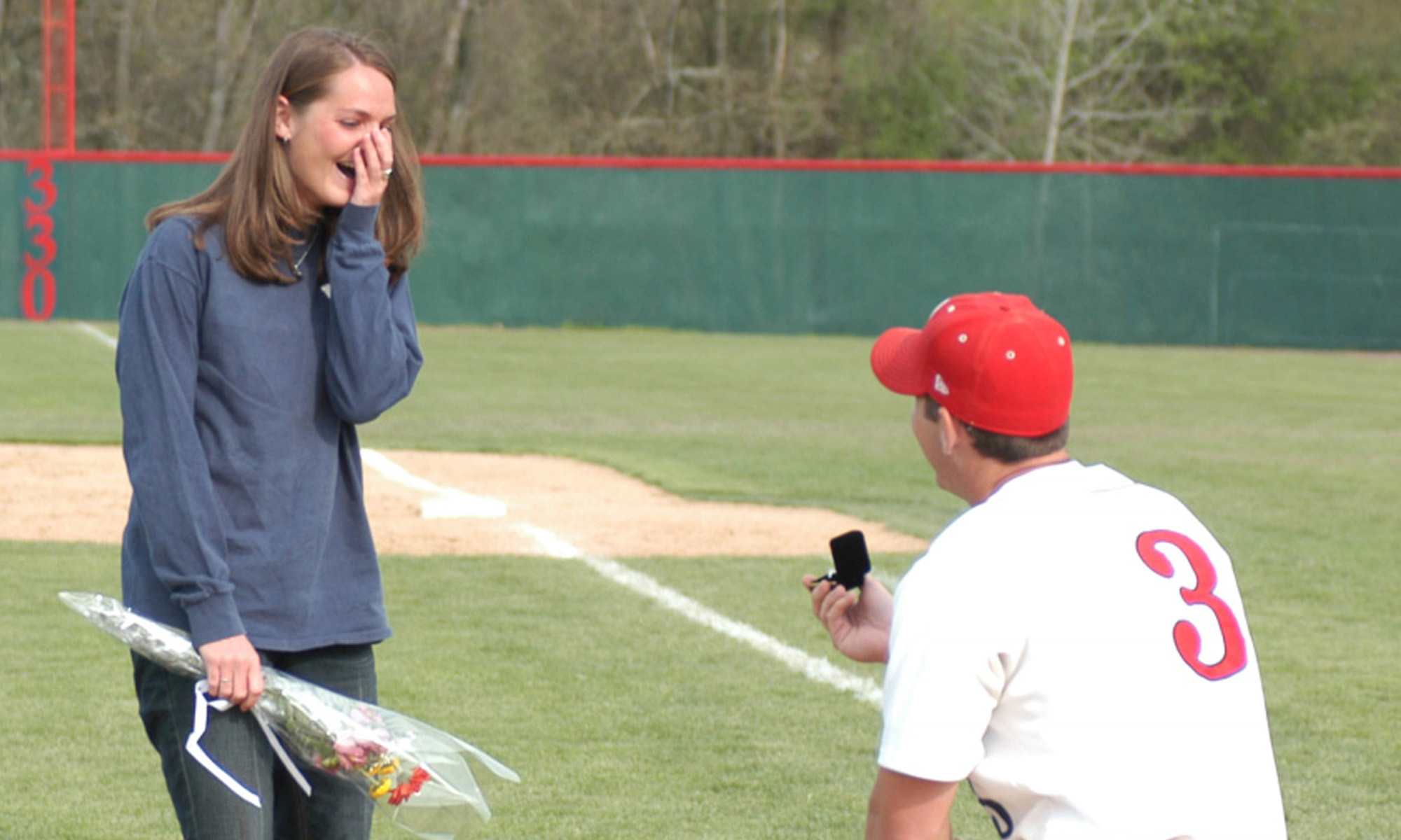 A baseball player in uniform kneeling and offering a wedding ring to a woman standing on a baseball field holding flowers.