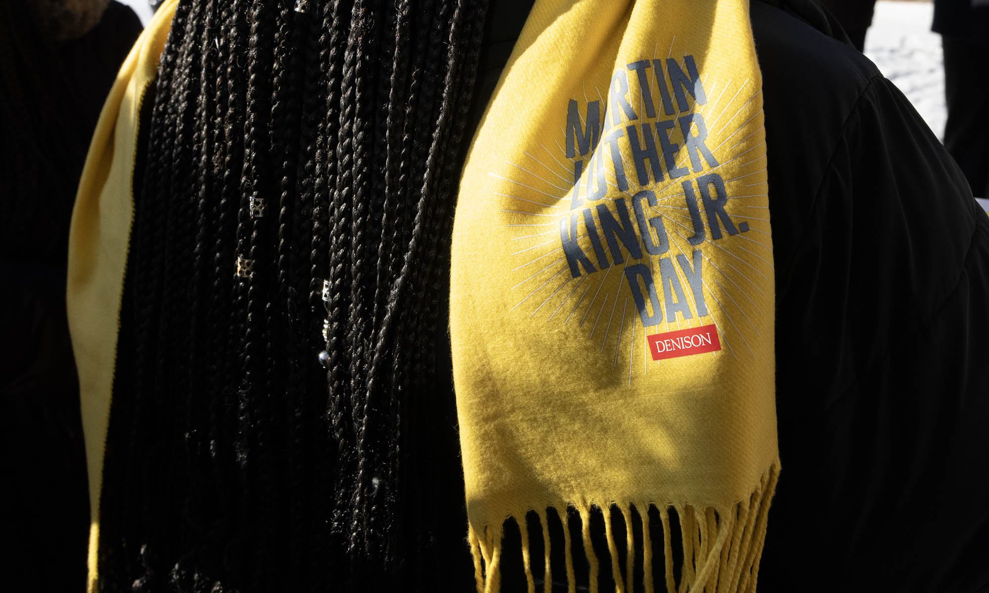 Close-up photo of a bright yellow scarf seen draped over a shoulder next to microbraids. The scarf reads Martin Luther King Jr Day Denison.