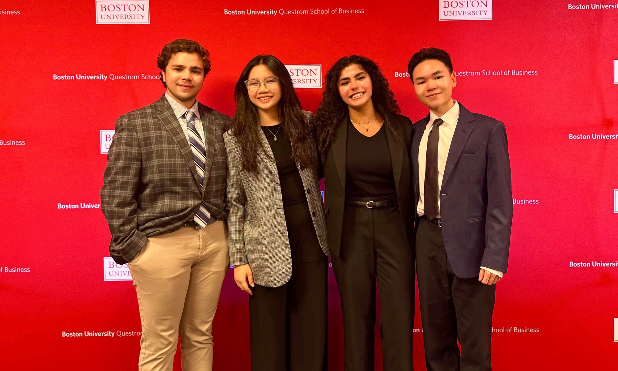 Four Denison students in business attire stand in front of a large red backdrop dotted with Boston University and BU Questrom School of Business logos ahead of their competition.