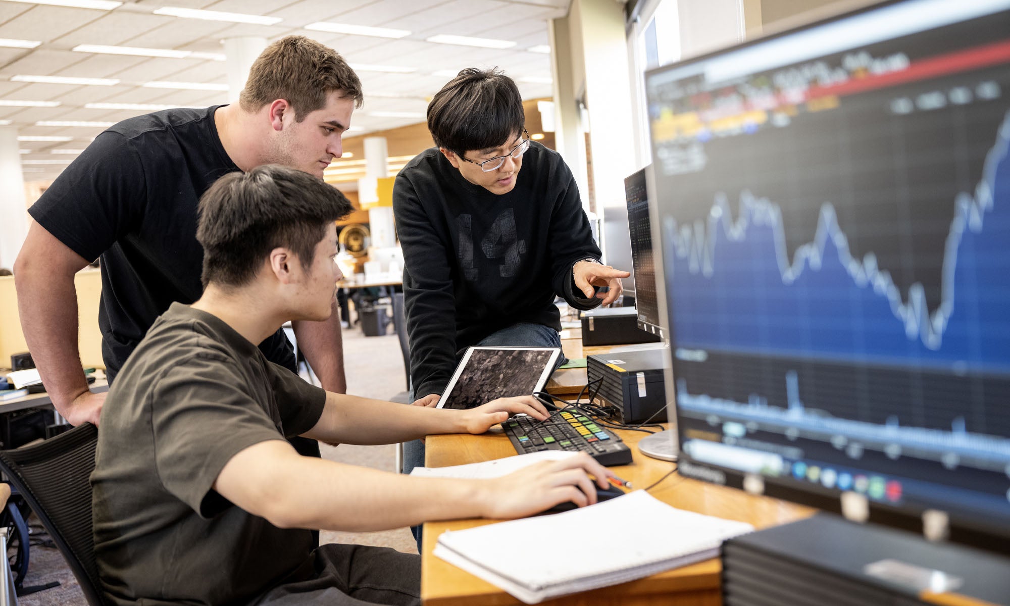 Three people in a computer lab look at a monitor together as one person points to an unseen data point; another monitor displaying stock market graph sits slightly out-of-focus in the foreground..