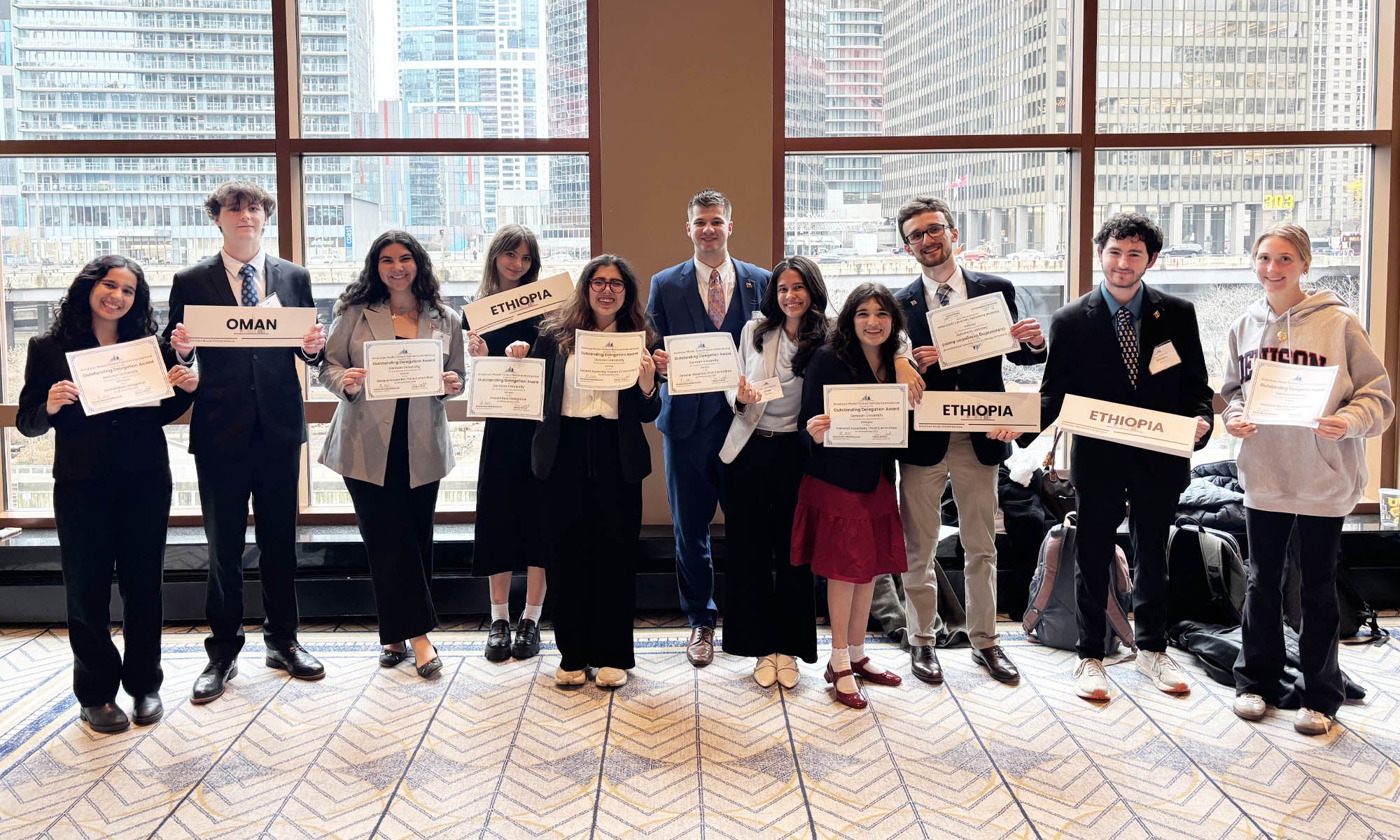 Eleven college students in business attire stand in front of large windows at a conference center, a view of an urban city center behind them, holding certificates and signs displaying the names of countries Oman and Ethopia.