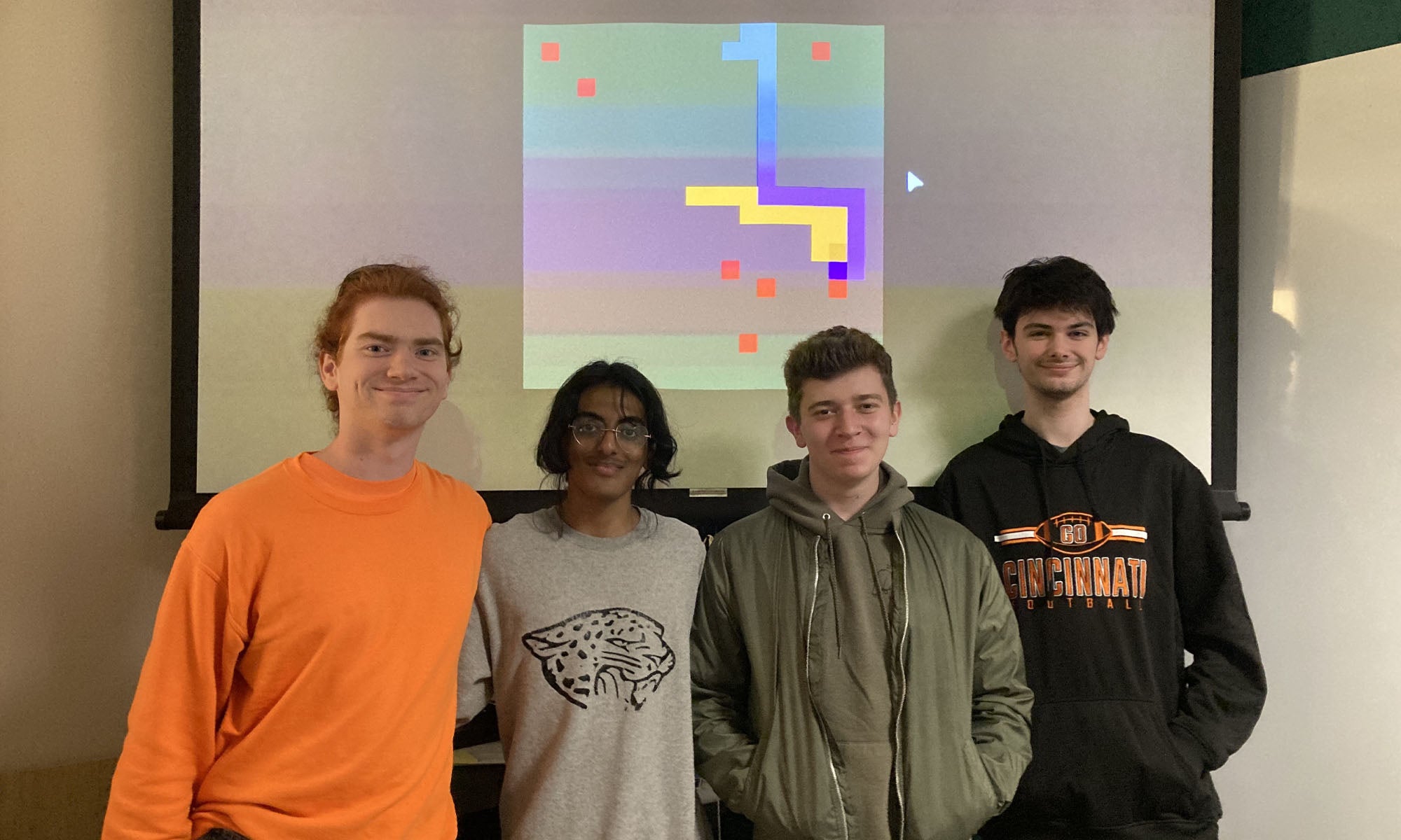 Four college students stand in front of a projector screen displaying a video game image resembling the classic computer "snake" game.