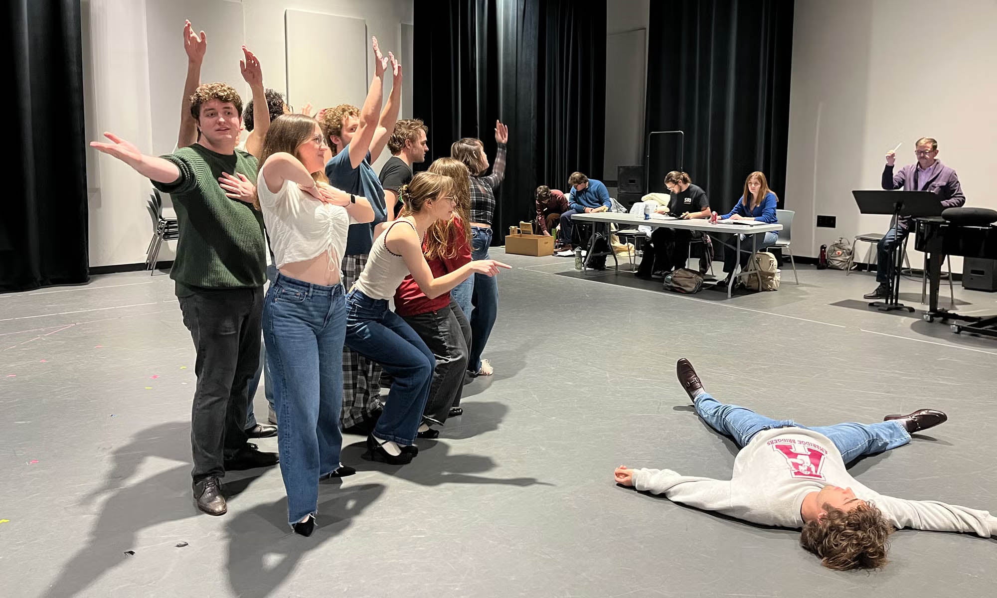 A group of actors rehearsing strike a pose during a choreographed number while the technical team and director watch from the wings.