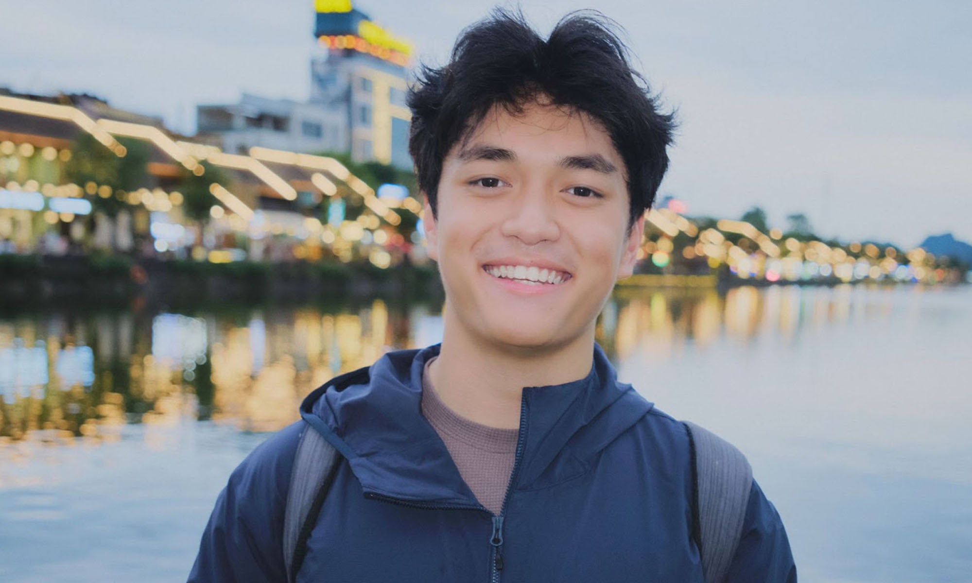 A young man with dark hair and a blue jacket smiles at the camera, standing by a waterfront with buildings and lights in the background.