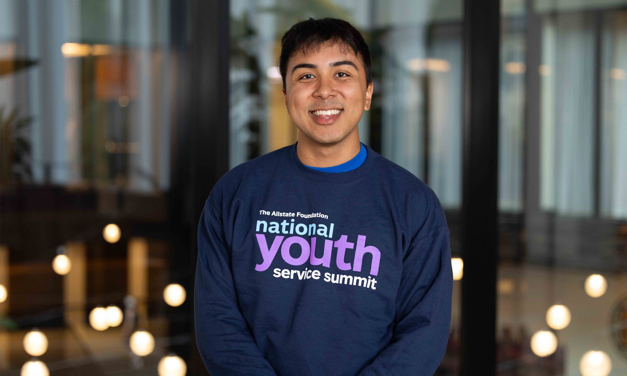 Photo of Yahir Fernandez-Alvarez ’26 wearing a National Youth Service Summit shirt, standing in front of a large glass wall inside a modern building.