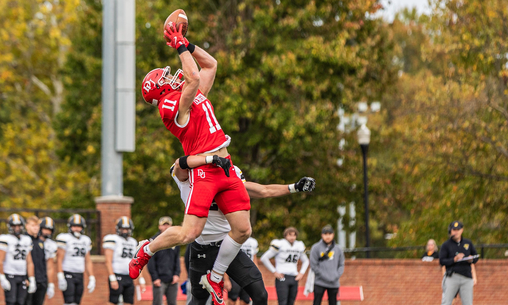 A football player in a red uniform jumps to catch a pass while a defender in a white and black uniform attempts to block him. Other players and trees are visible in the background.