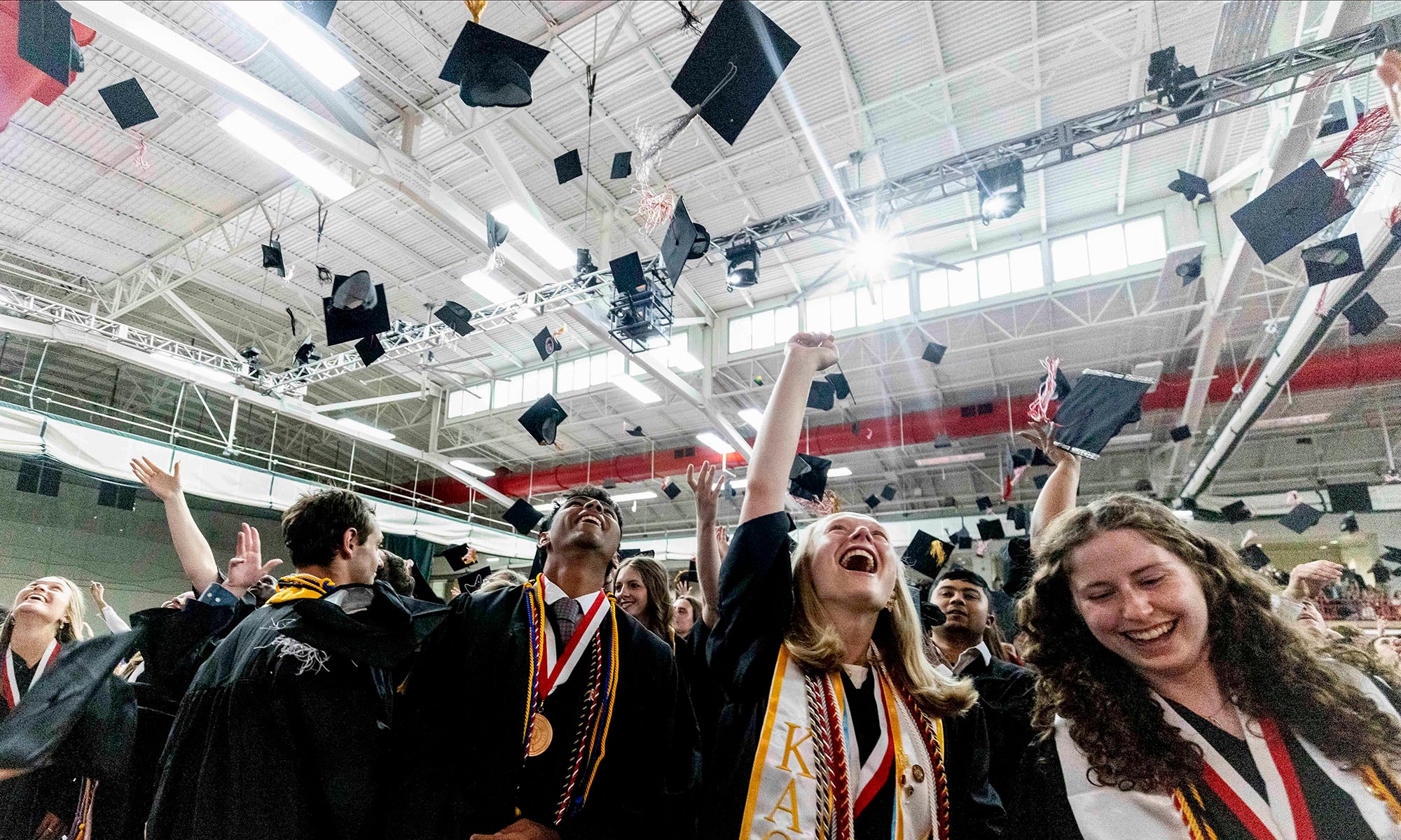 A large group of college graduates in caps and gowns toss their caps in the air in celebration in a large fieldhouse during a graduation ceremony.