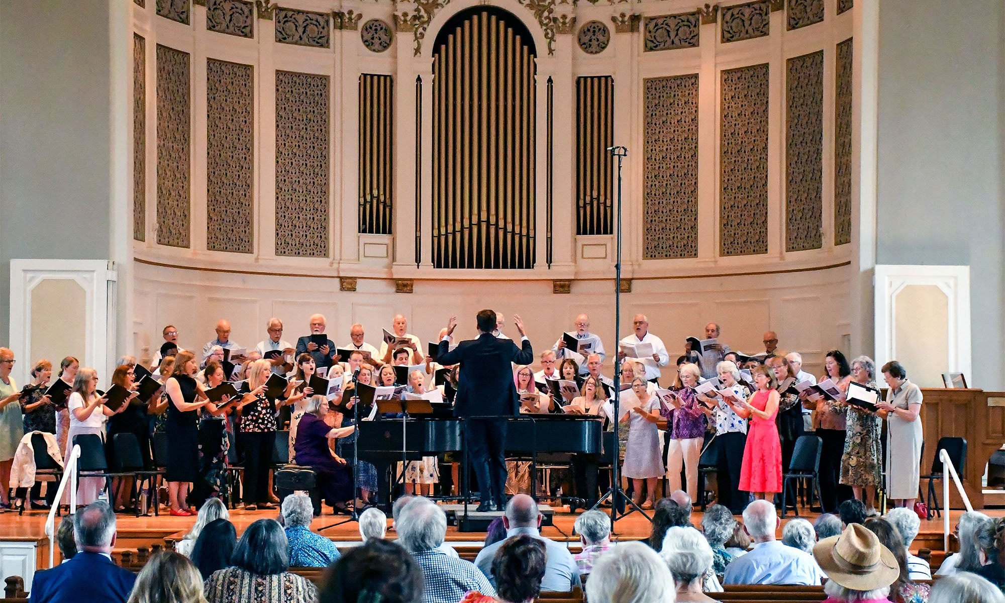 A choir and musicians perform on stage in a church or concert hall, directed by a conductor. The audience is seated in rows facing the performers. The background features a large organ and decorative architectural details.