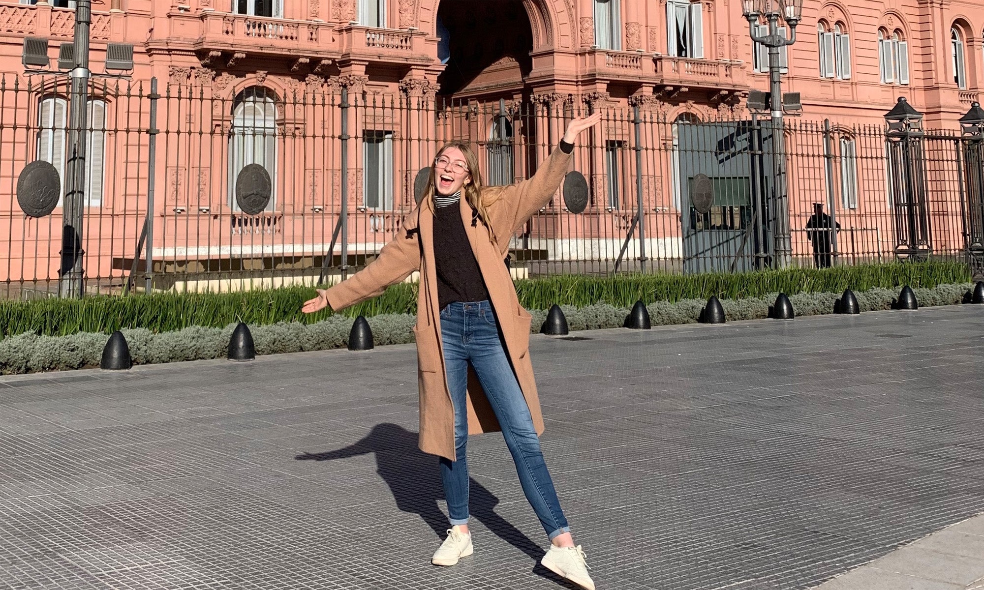 A study abroad student in a tan coat and jeans stands smiling with arms outstretched in front of a large pink historic building with black iron fencing.