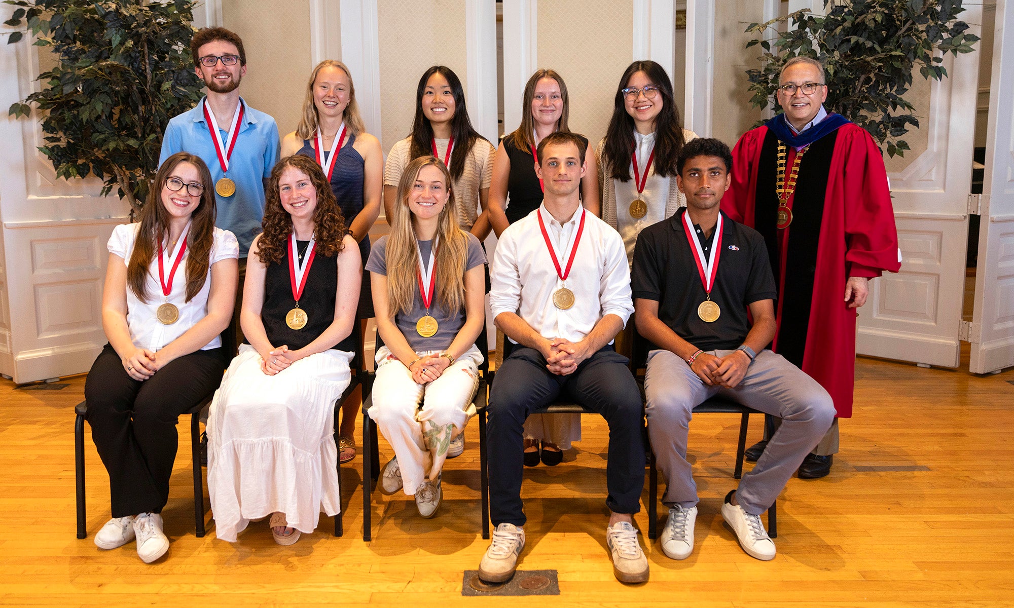 A group of ten Denison students, all 2026 President's medalists with medals, and President Adam Weinberg in academic regalia pose indoors, with plants and wood flooring in the background.