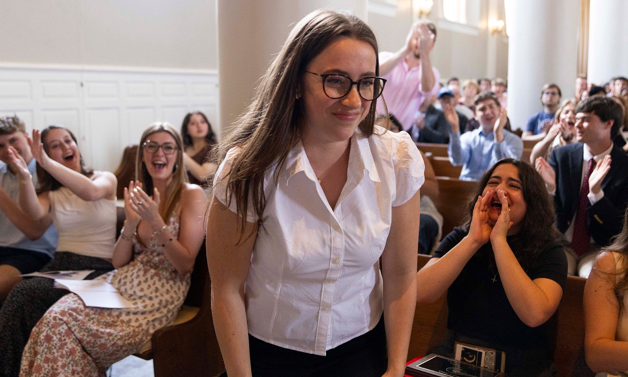 A young woman in a white shirt stands smiling as people around her seated in an auditorium applaud during the Academic Awards Convocation.