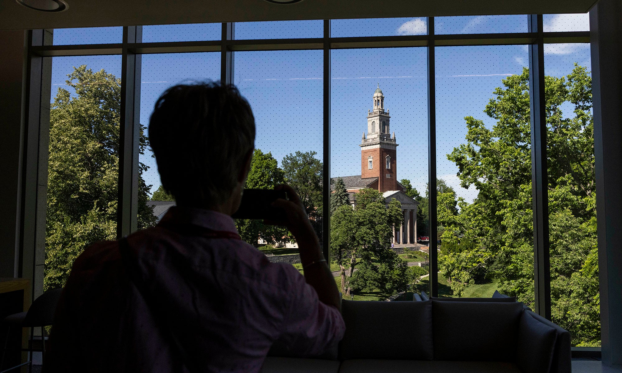 A person takes a photo of a college campus clock tower through large indoor windows on a sunny day.