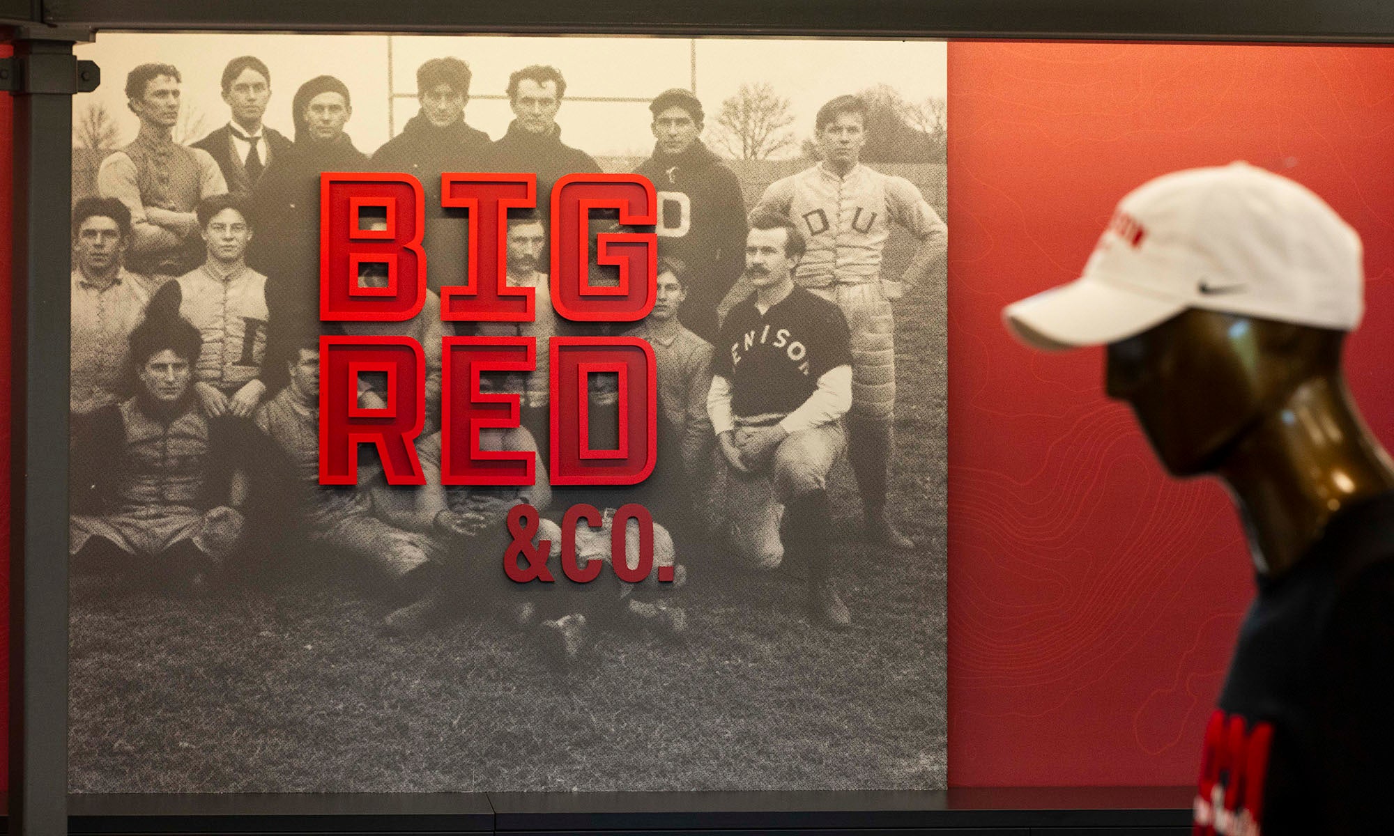 A mannequin in a white cap stands beside a vintage black-and-white team photo with large red letters reading BIG RED on the wall.