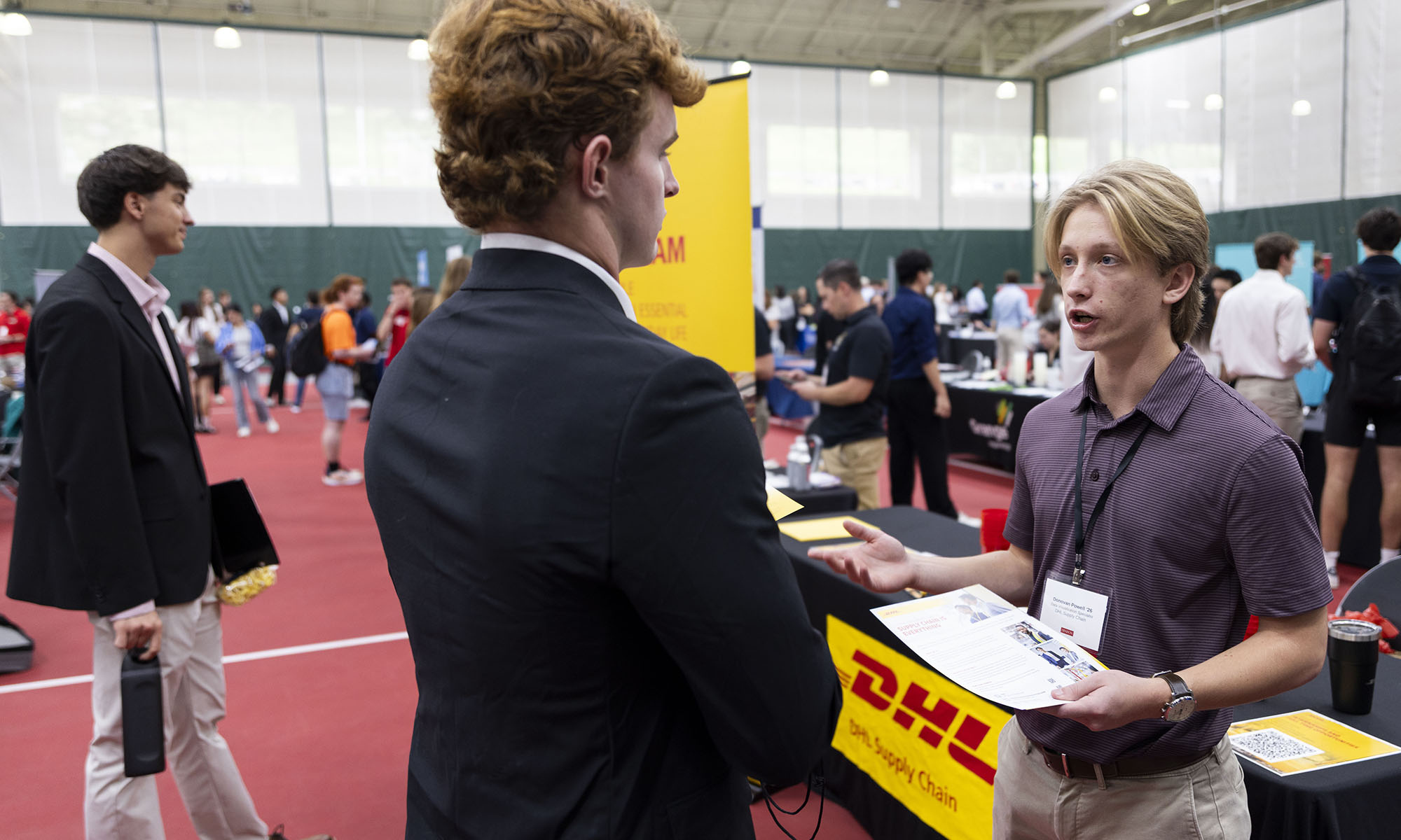 Two people speak at a career fair booth with a visible DHL sign, while others interact in the background inside a large gymnasium.