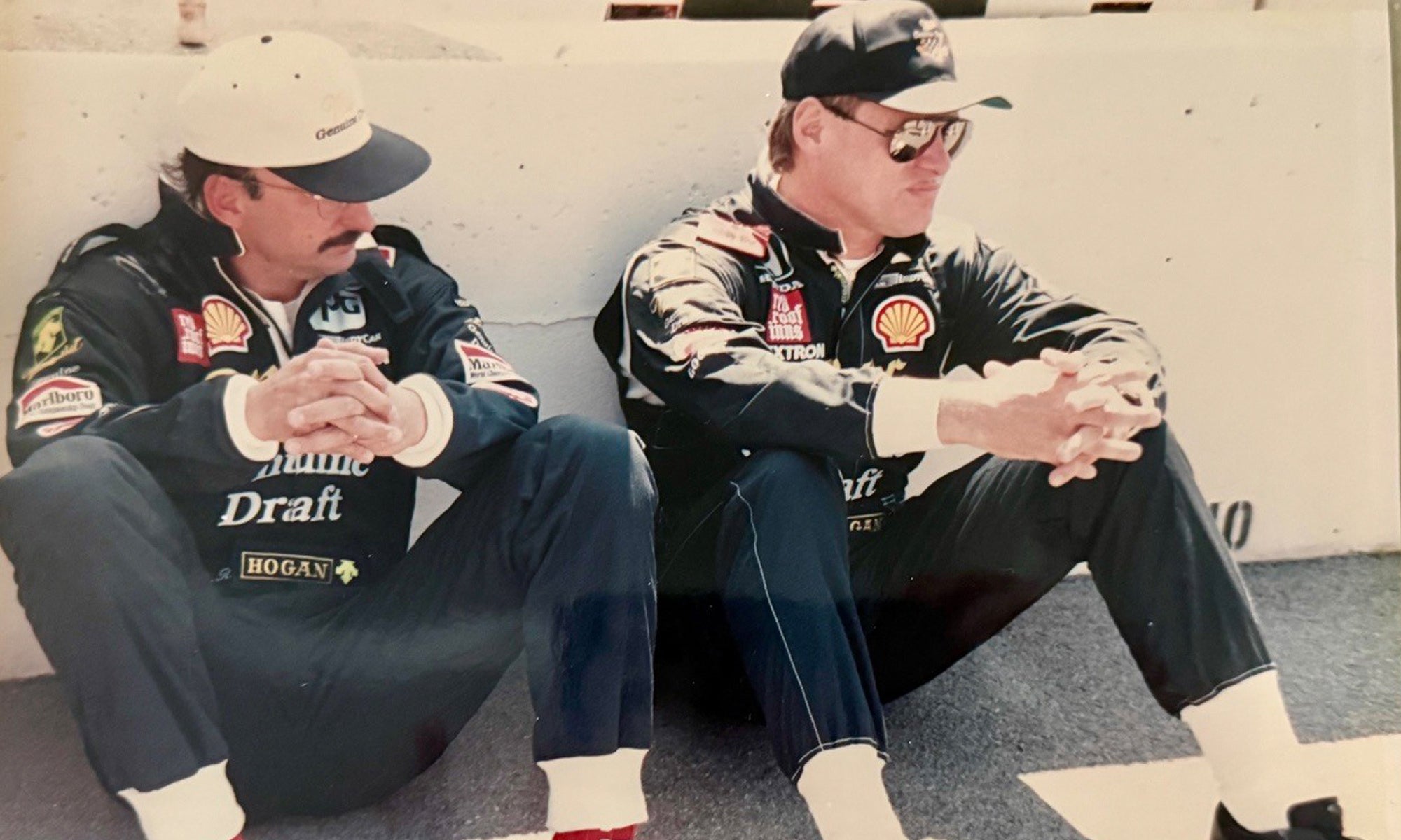 Two men in racing uniforms and caps sit on the ground against a wall, looking forward with hands clasped over their knees.