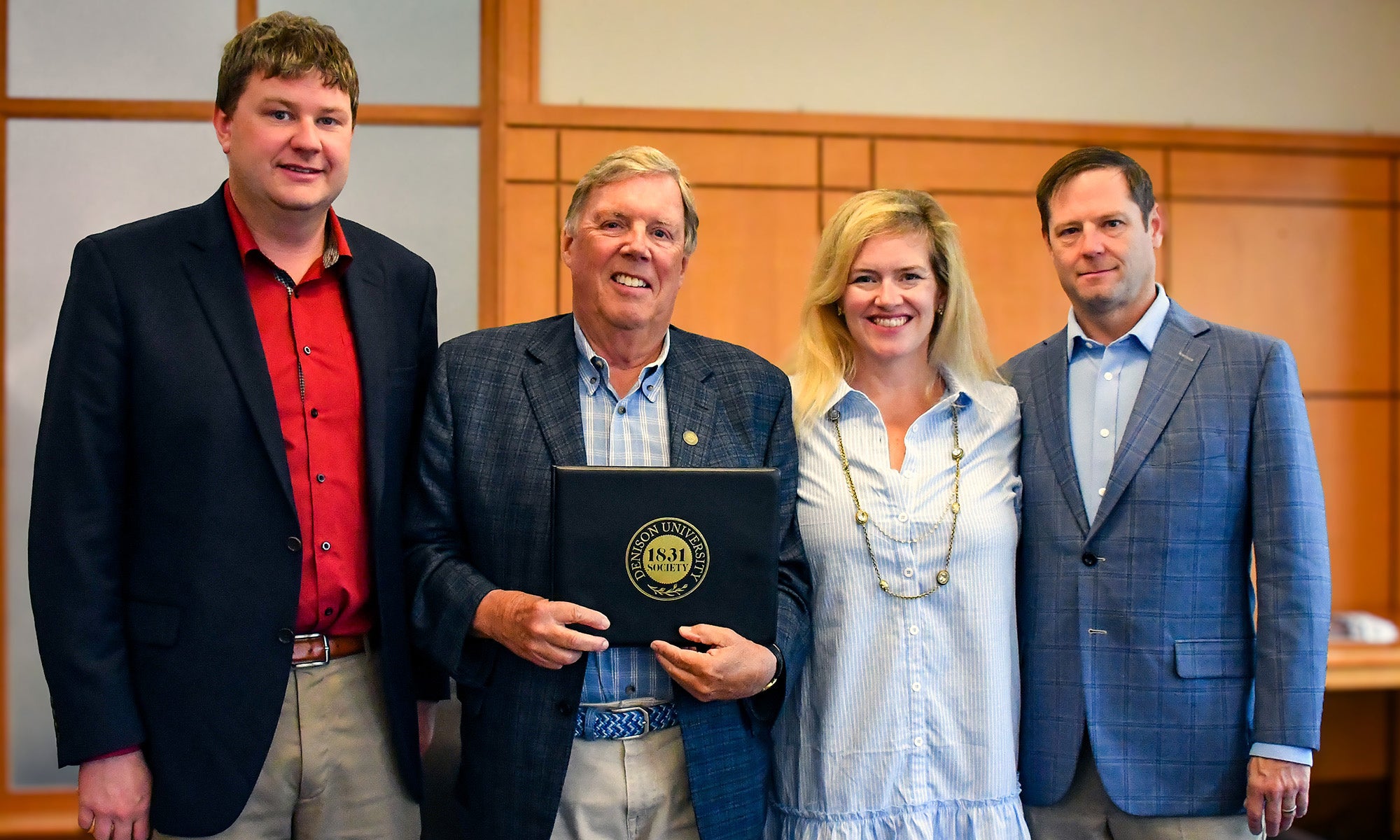 Four people stand indoors; the man second from left holds a certificate and smiles at the camera, while the others stand close by, all dressed in business or business-casual attire.