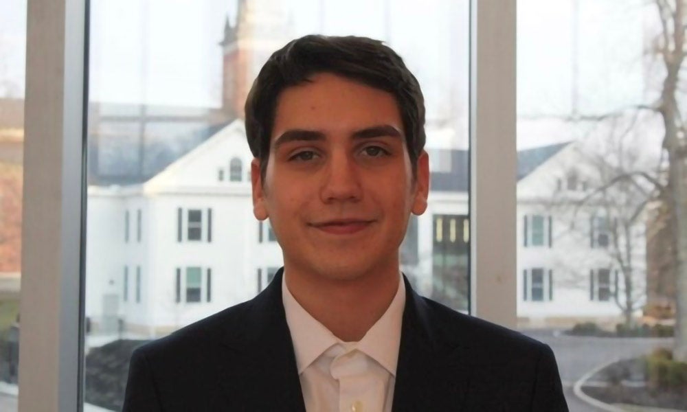  Max Levine ’26 wearing a dark suit jacket and white button-up shirt stands in front of a large window; Denison's Reese House and Swasey Chapel viewed in the background.