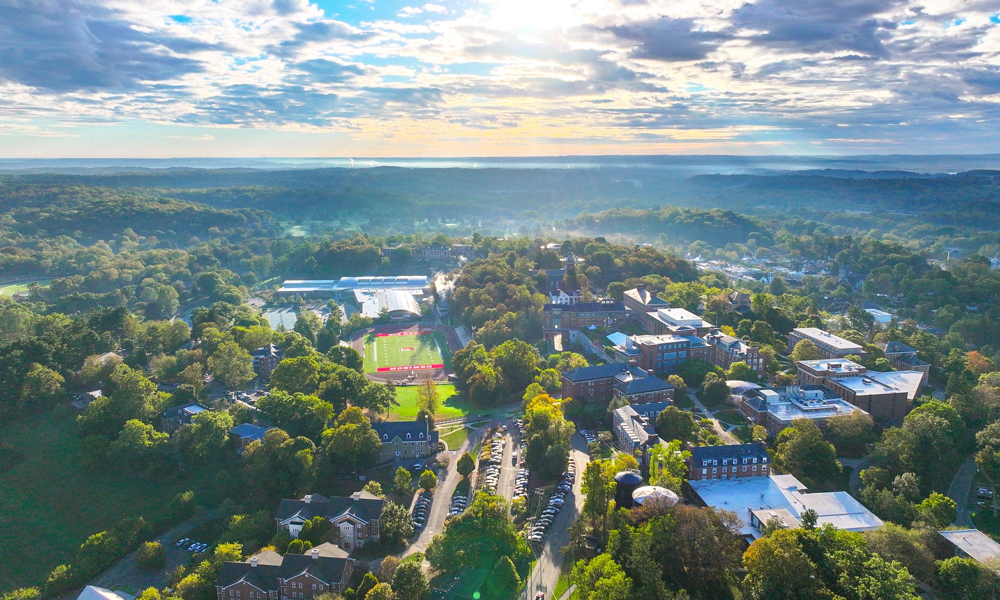 https://denison.eduAerial view of a college campus with multiple buildings, sports fields, and surrounding trees under a partly cloudy sky.