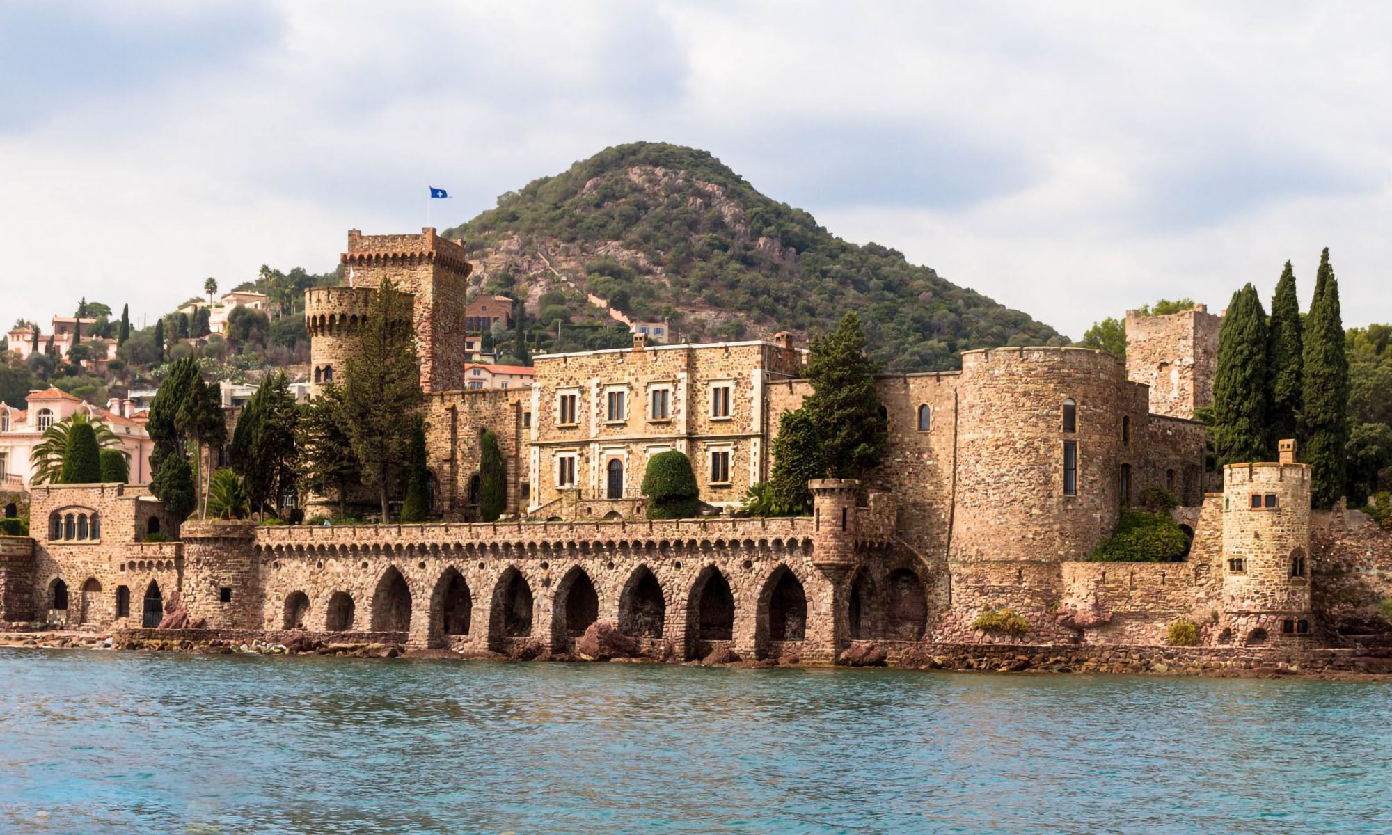 A historic stone castle with arched walkways and towers sits beside a lake, with a wooded hill rising in the background under a cloudy sky.