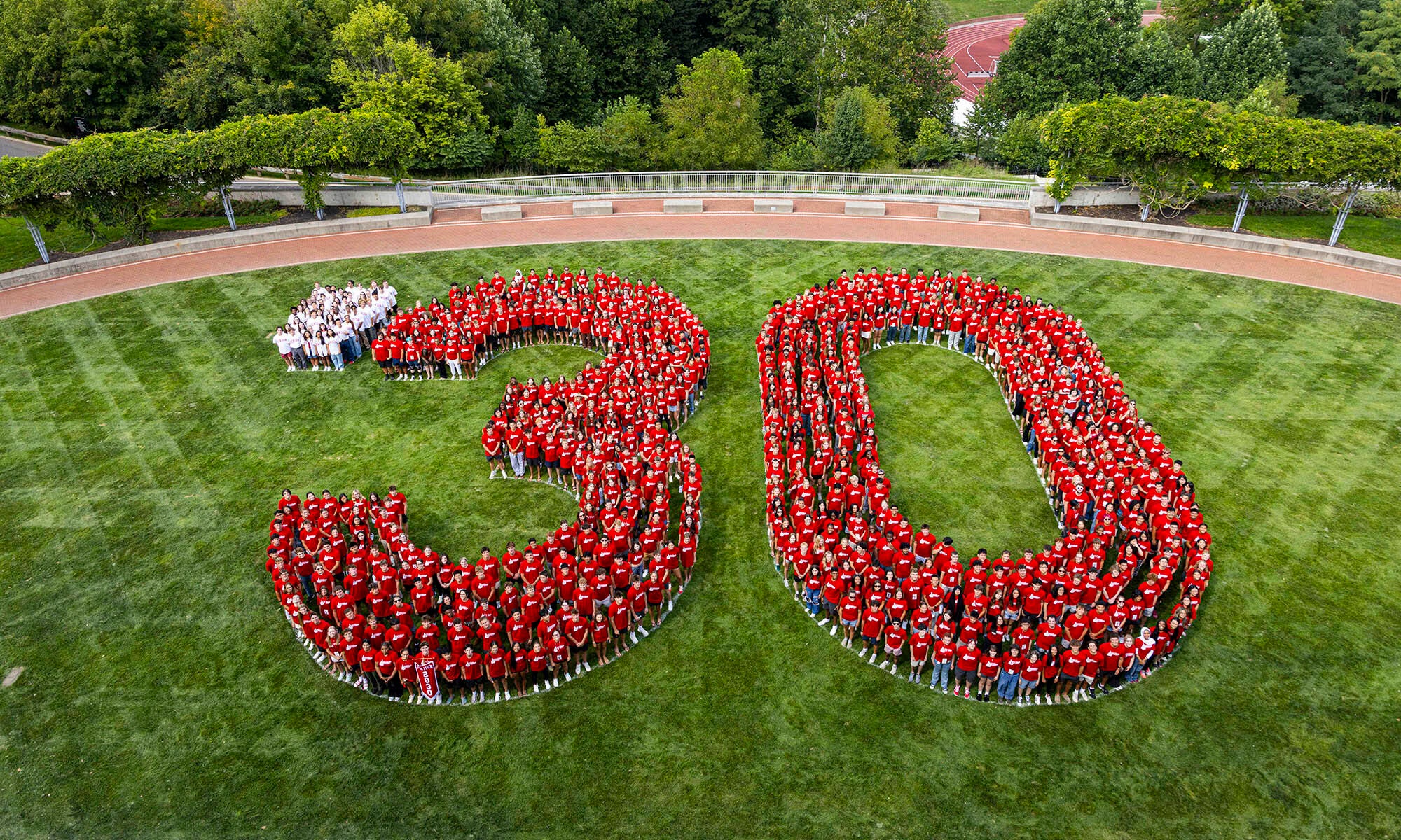 A large group of people wearing red shirts stand on grass arranged to form the number 30, with a few people in white shirts forming a dot before the 3. Trees and a path are in the background.