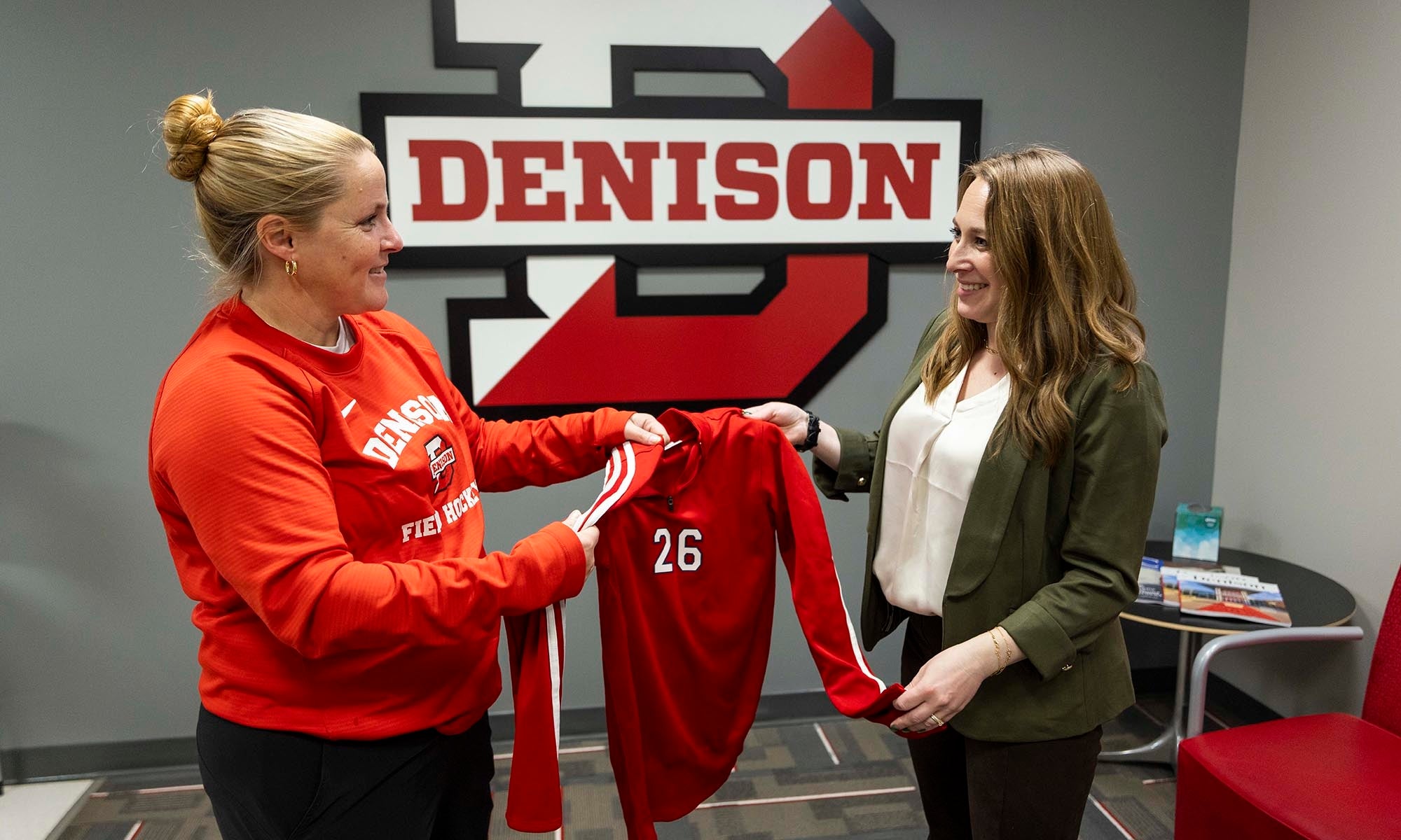 Two women stand indoors in front of a large Denison University logo. One hands a red sports jersey with the number 26 on it to the other. Both are smiling.