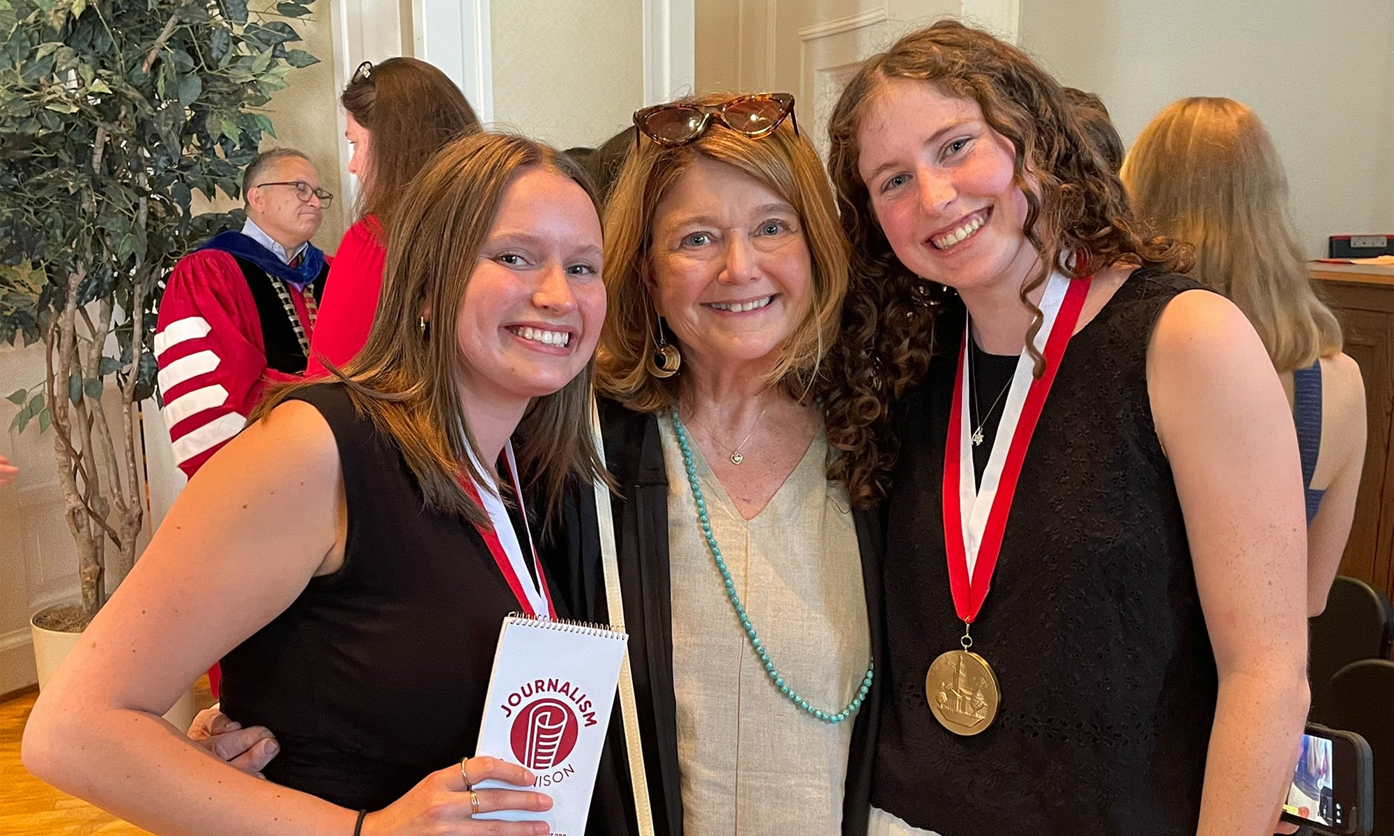 Three women stand indoors, smiling at the camera. Two younger women wear medals on red and white ribbons around their necks, and the other woman stands between them. Other people are visible in the background.