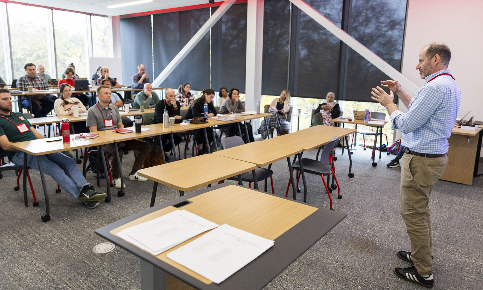 A man stands at the front of a classroom, gesturing while speaking to a group of seated adults.
