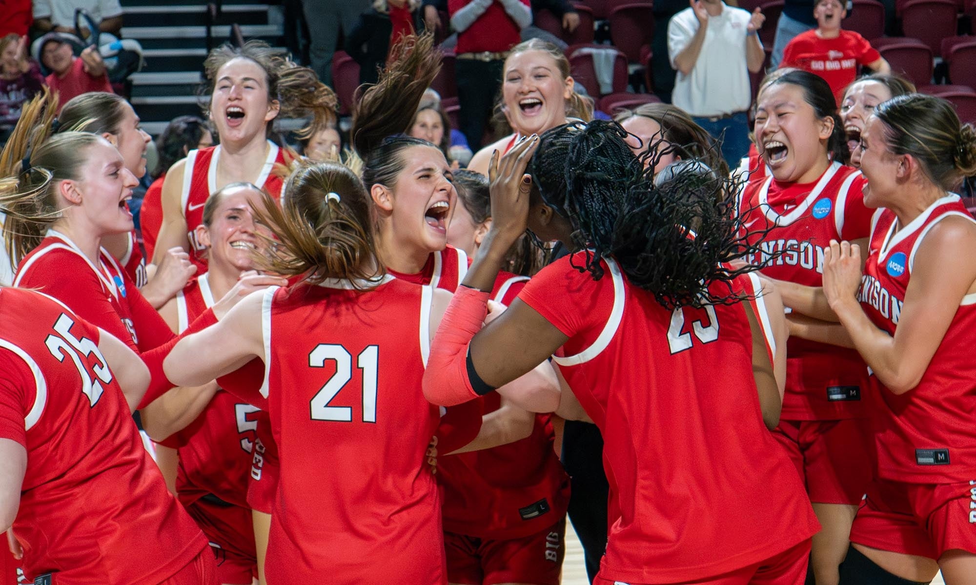 A womens basketball team in red uniforms celebrates together on the court, laughing and cheering. Player number 21 is visible in the center of the group.