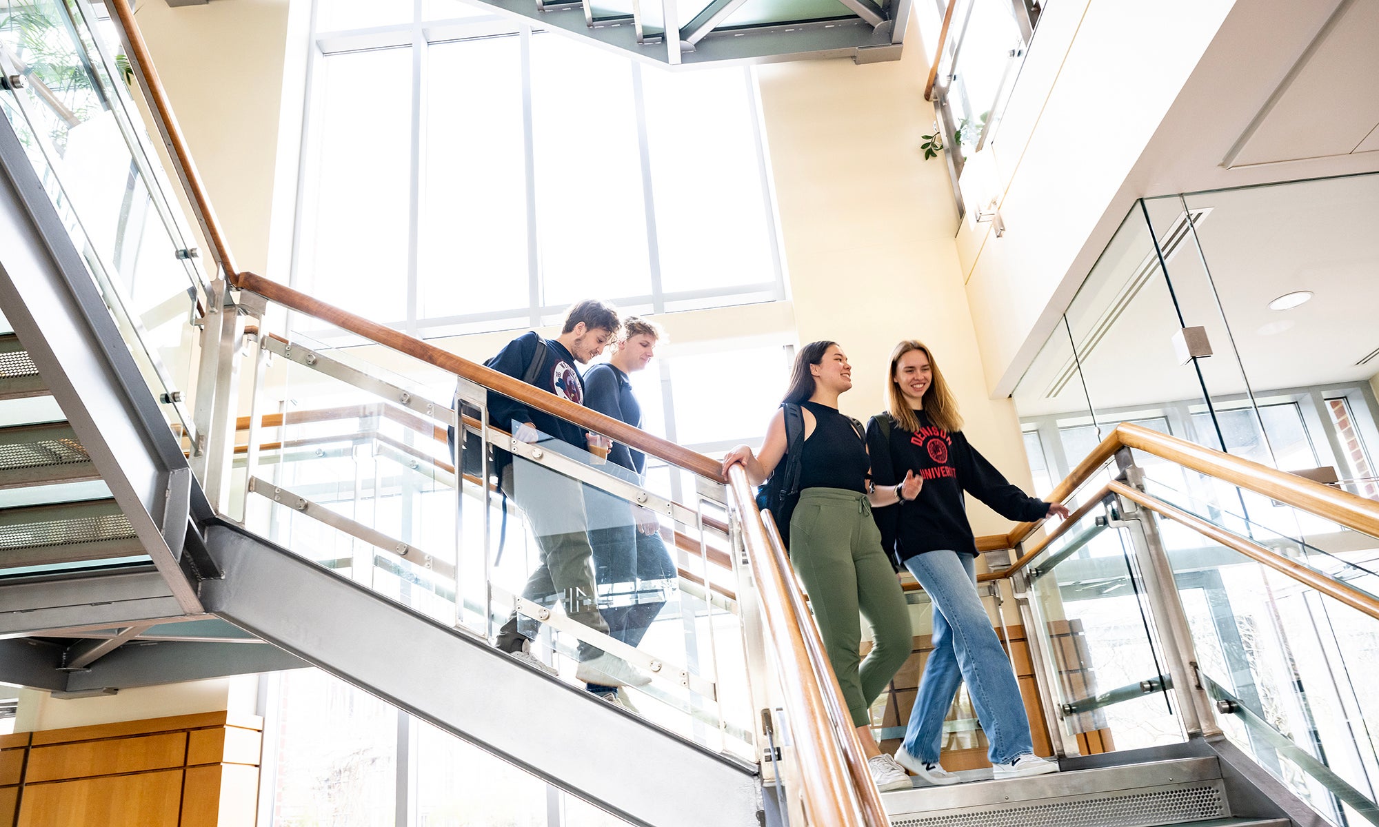 https://denison.eduFour people walk up and down a bright, modern staircase in a building with glass walls and metal railings.
