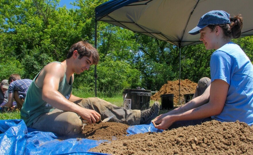 Two people sit on the ground under a canopy, sifting through dirt on a blue tarp, working on an outdoor excavation or archaeology project (Photo credit: Allie Vugrincic)