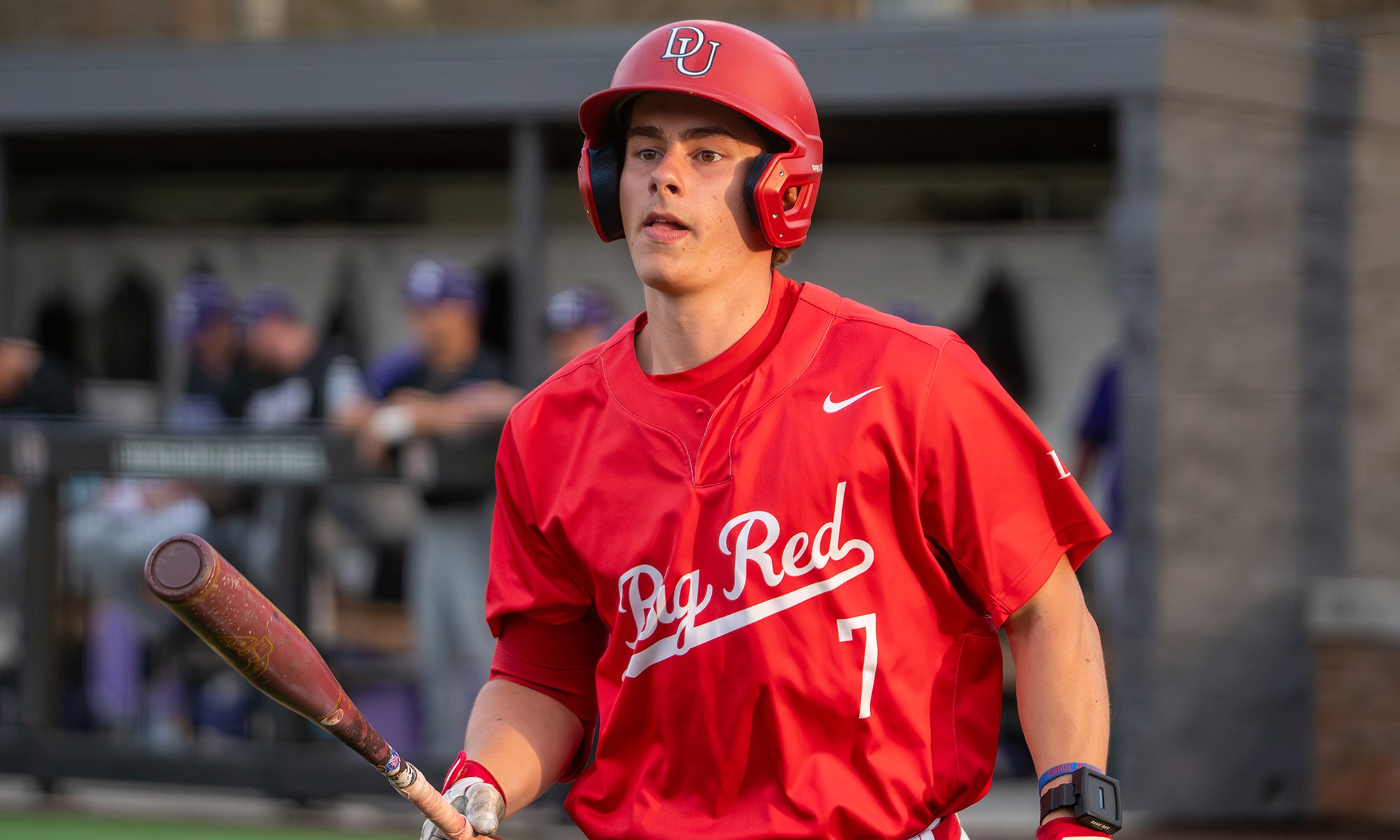 Baseball player in a red uniform and helmet holding a bat stands near the field, with a dugout and other players visible in the background.