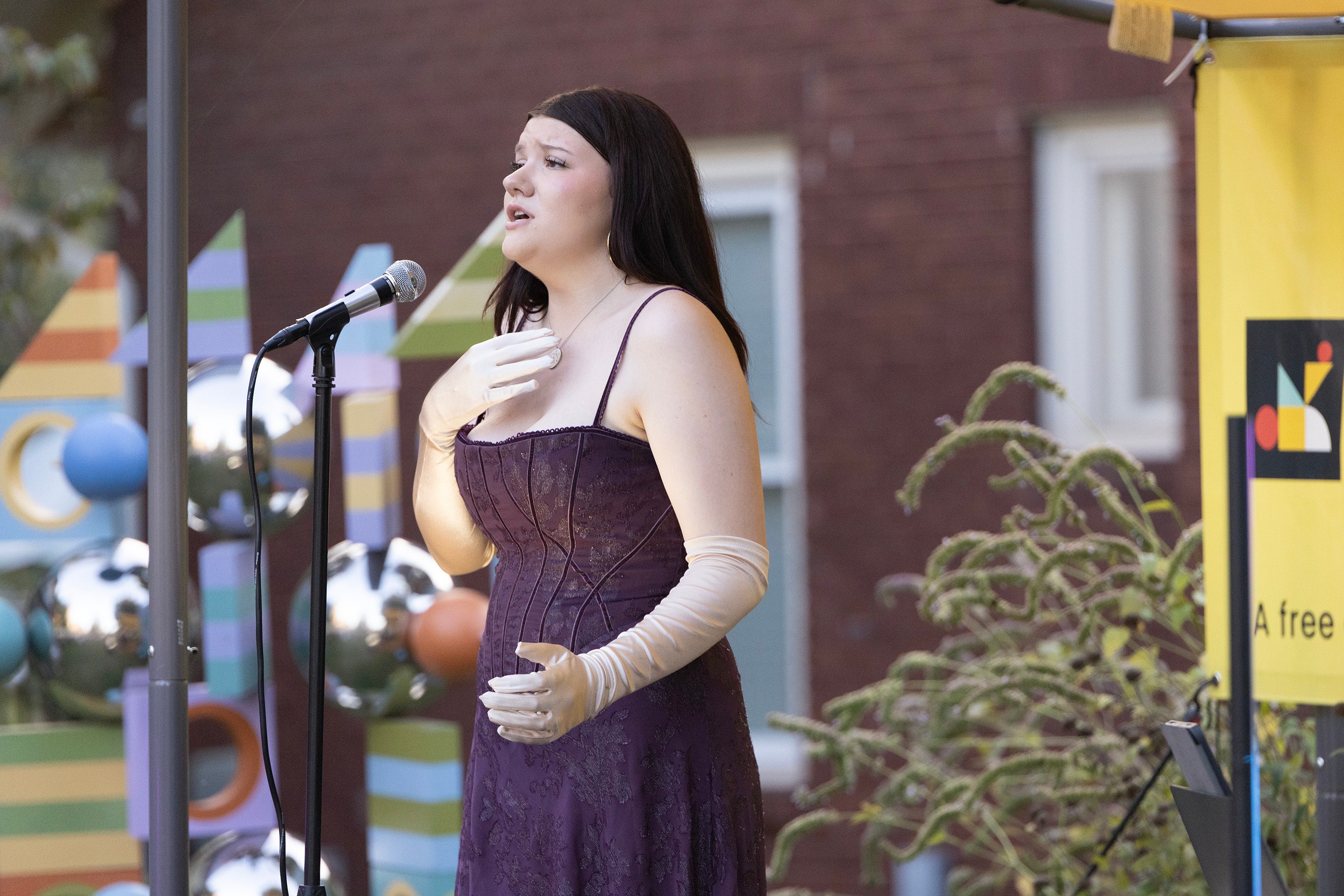 A woman with dark hair, wearing a purple sleeveless dress with straps, sings on an outdoor stage.