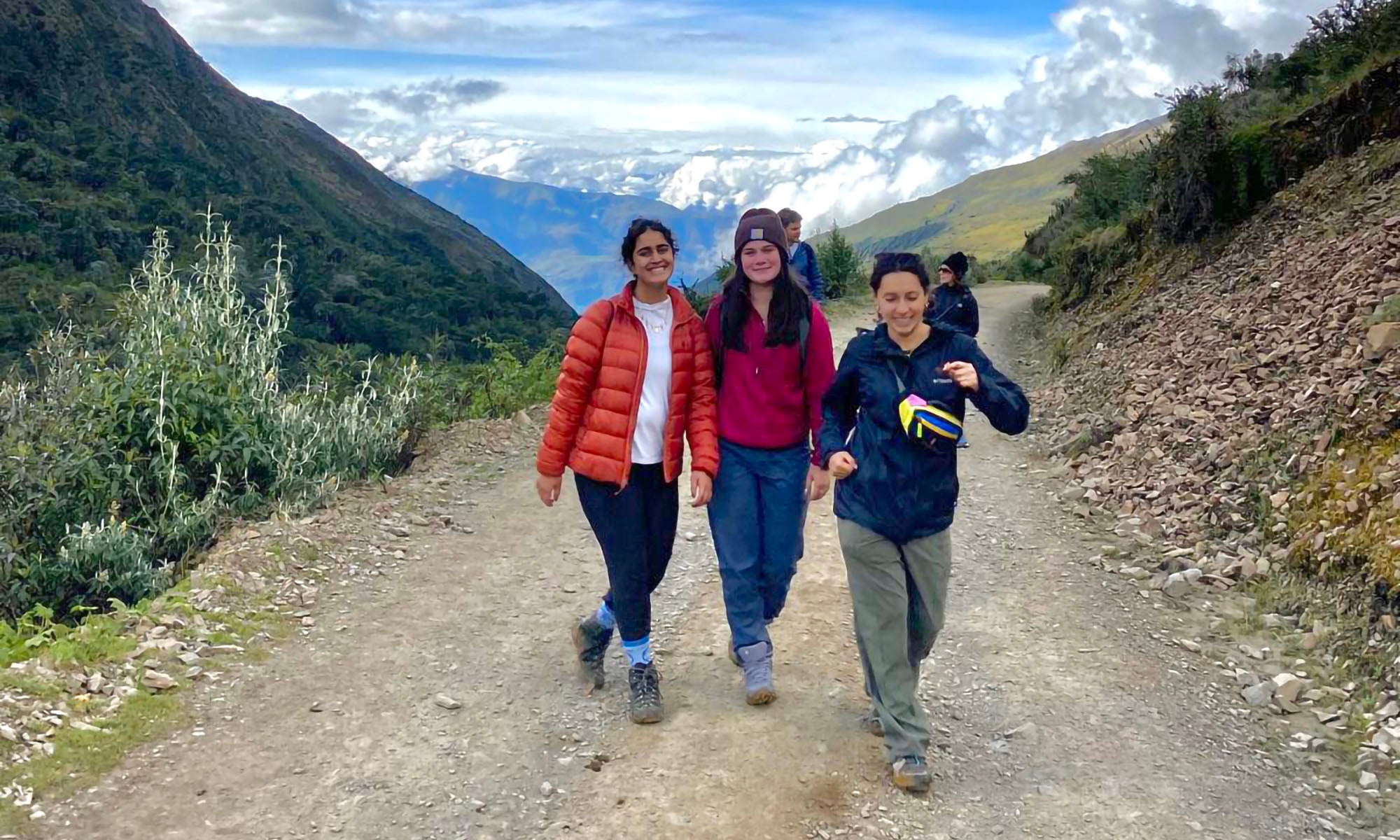Three people on a hiking on a high-altitude trail with snow-capped mountains and clouds in the distance behind them.