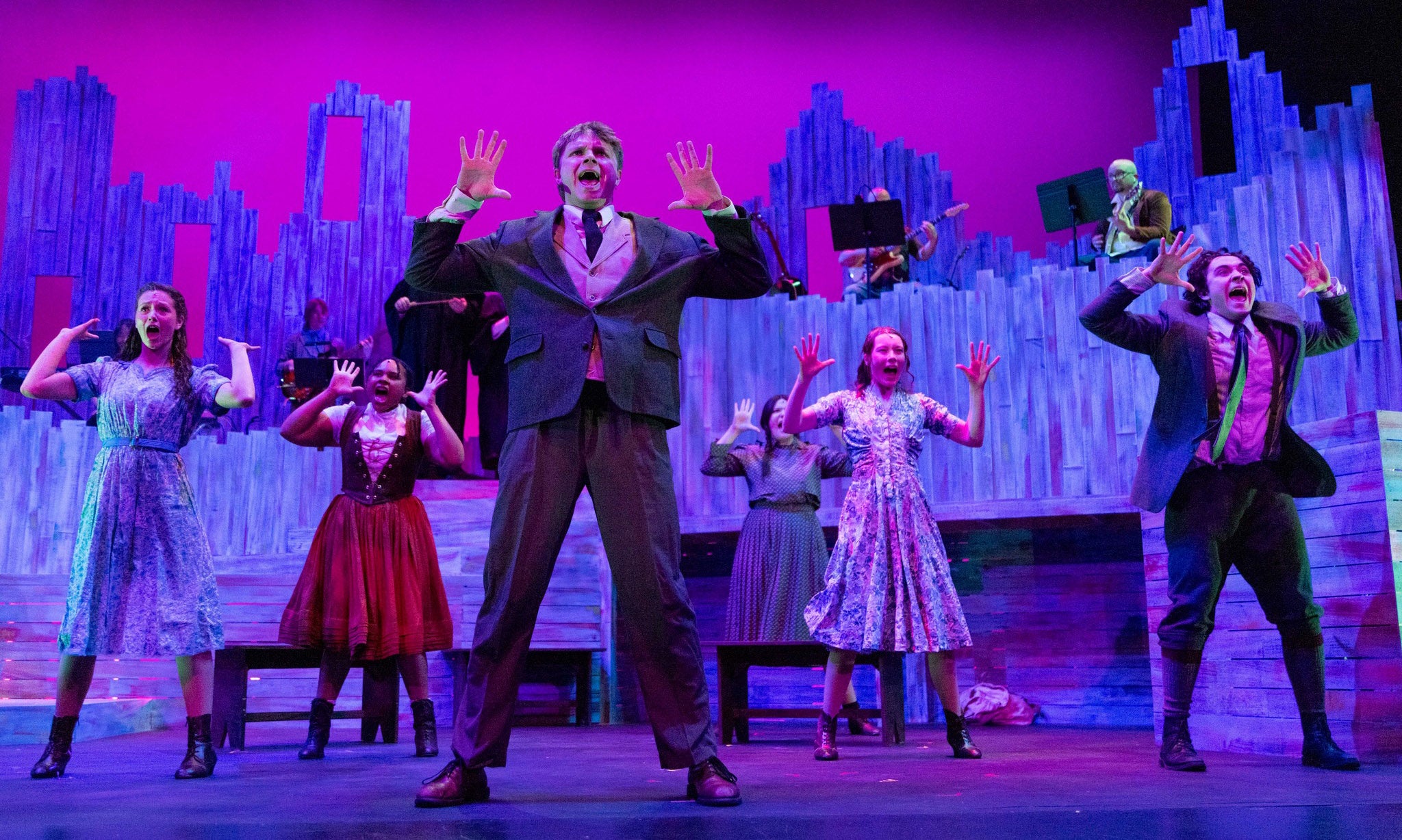 A group of performers on stage sing and gesture with their hands raised. They are dressed in formal and vintage-style costumes, with a colorful, abstract cityscape set and vibrant purple lighting in the background.