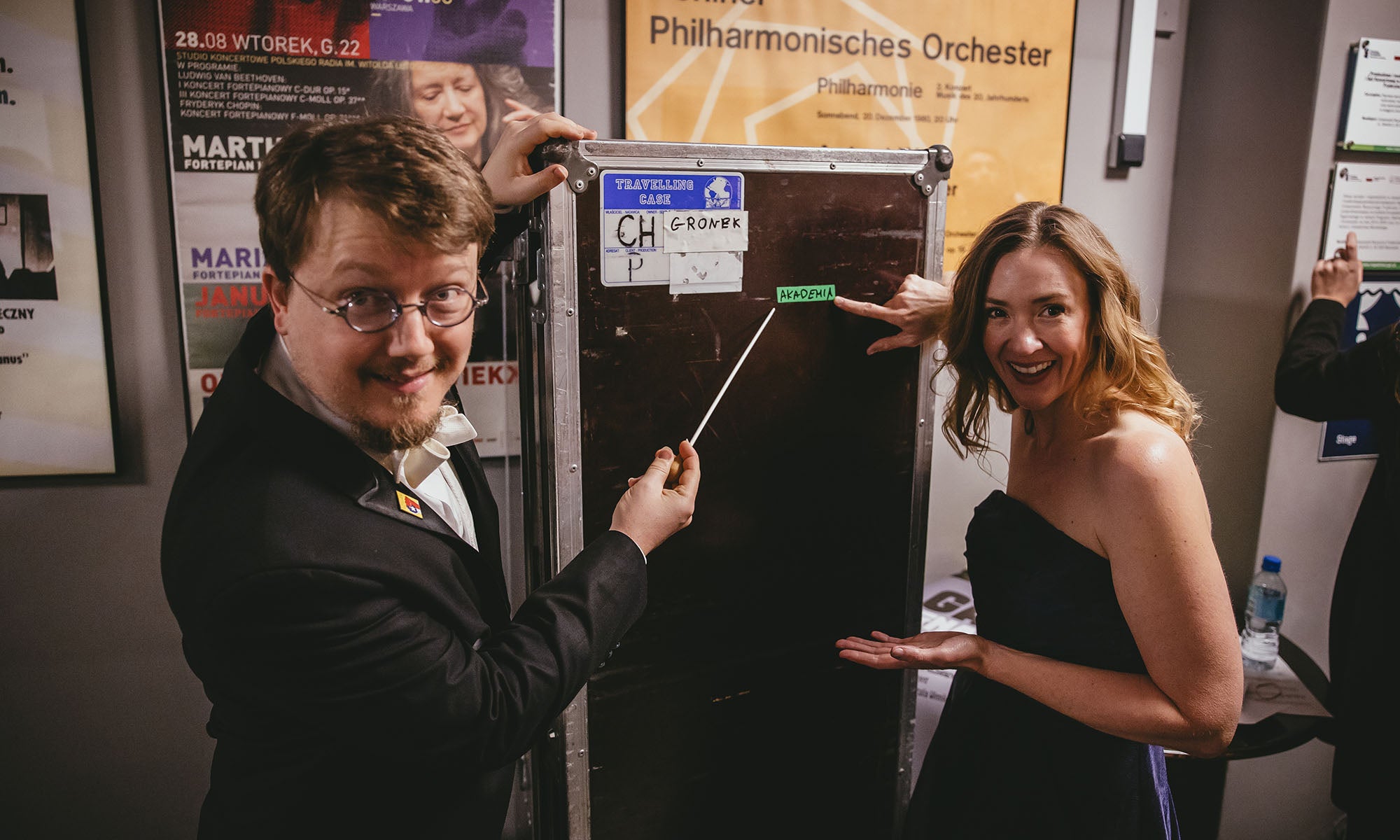 Two people in formal wear smile and point to a music storage case bearing a sticker that says "Akedemia."