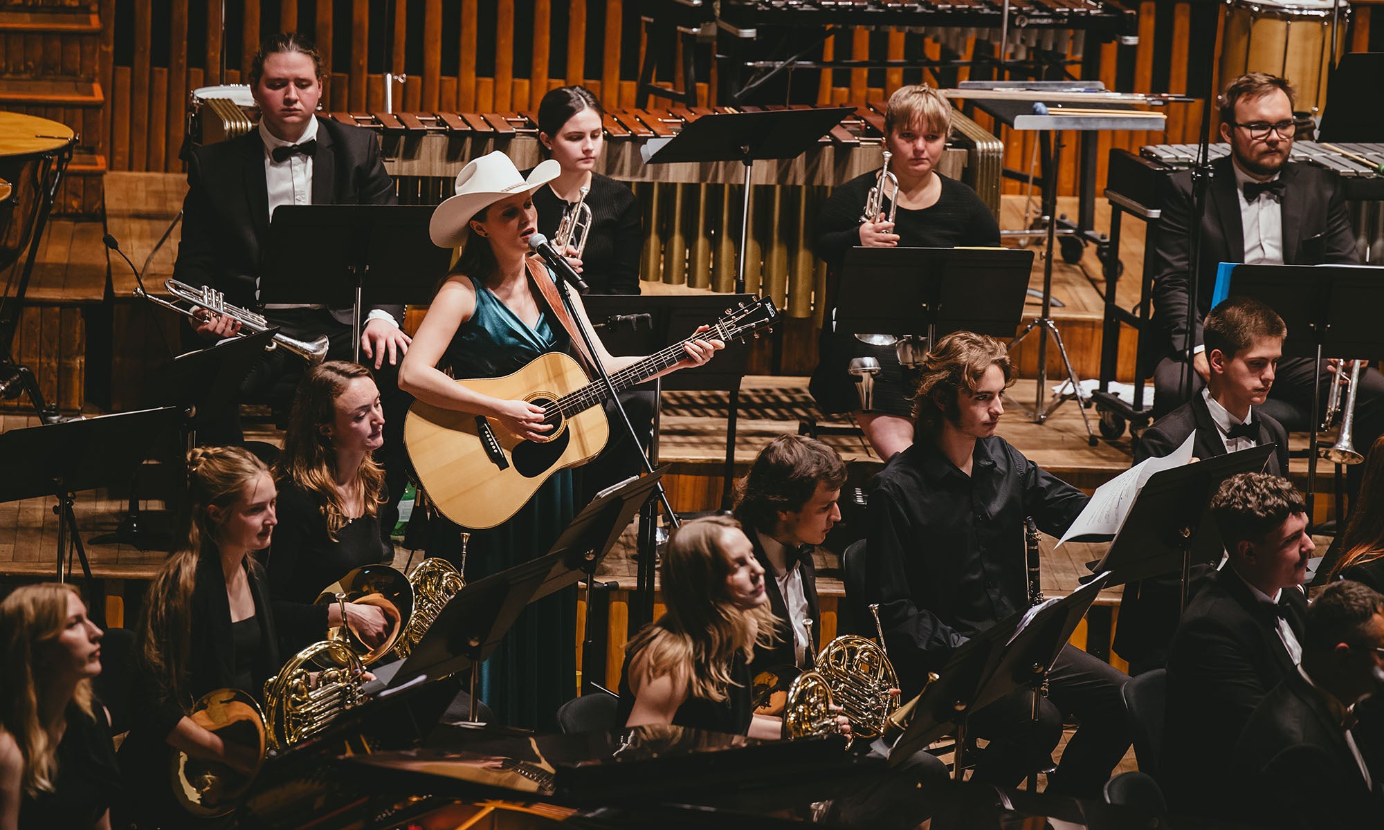 A woman in a white Stetson cowboy hat plays acoustic guitar and sings on stage with a seated orchestra of musicians holding various instruments.