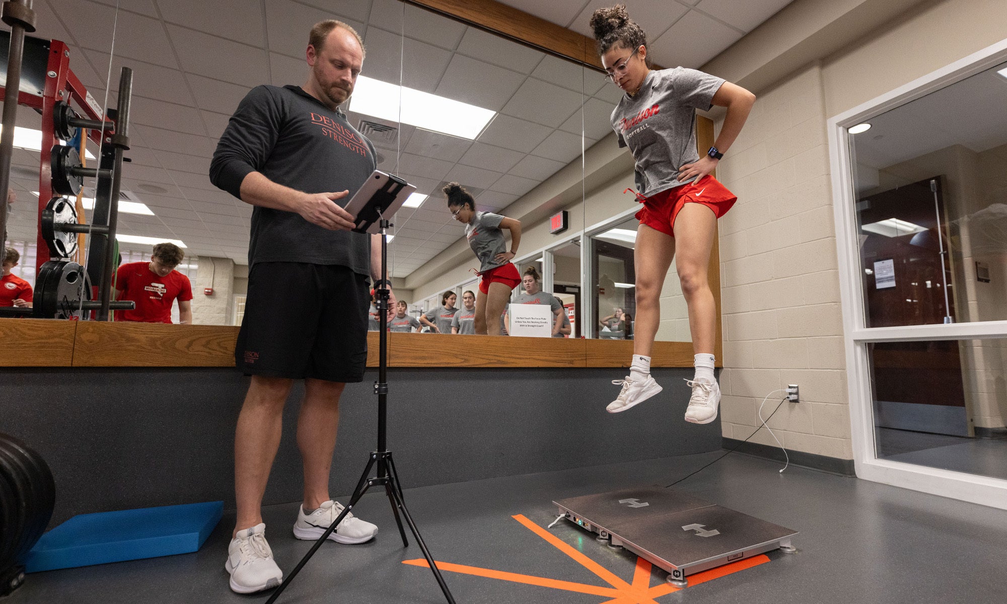A woman in athletic gear performs a vertical jump test while a man in sportswear records results on a tablet. The scene takes place in a gym with mirrors and exercise equipment visible in the background.