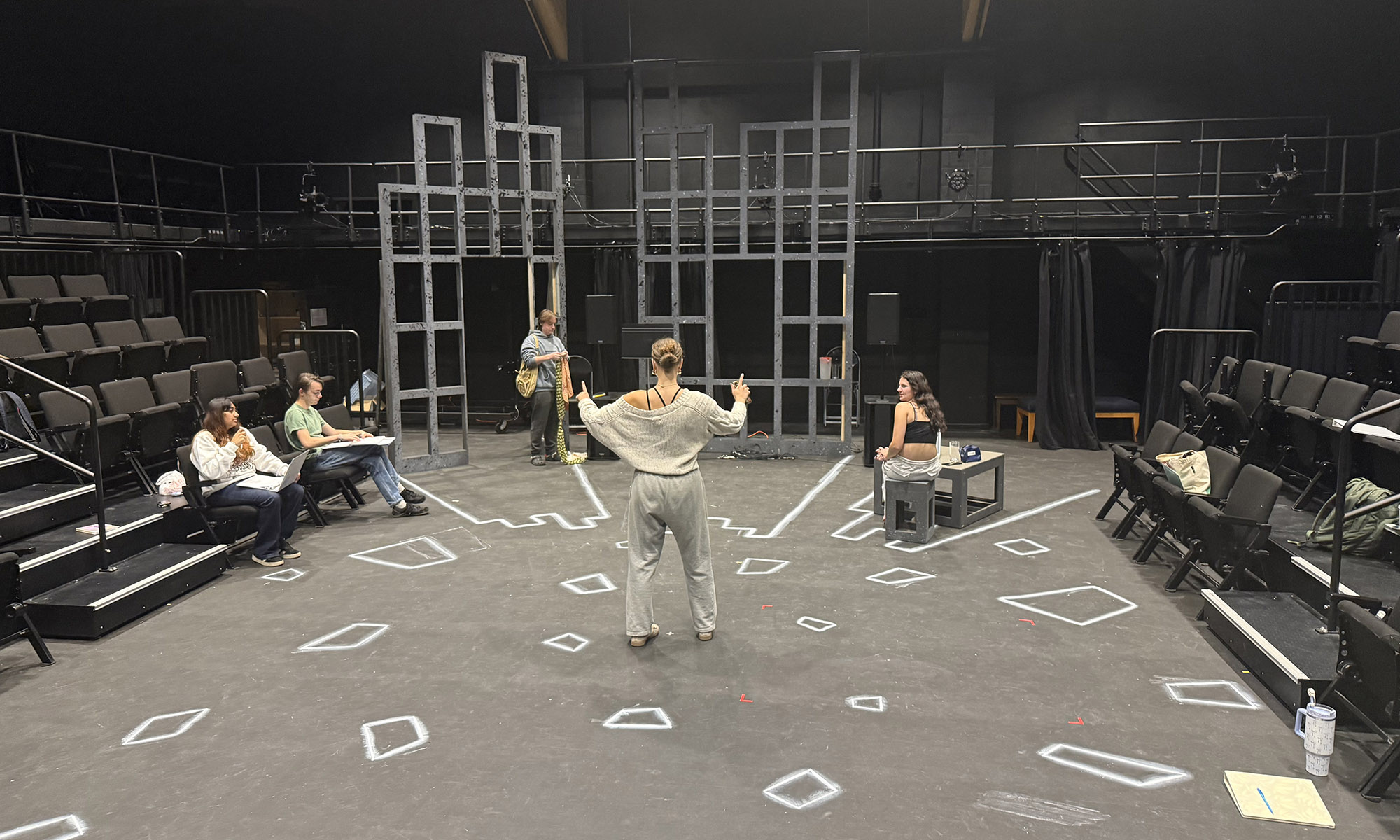 A person stands in the center of a theater stage marked with tape, facing four others seated or standing around the set, which includes geometric wooden frames and scattered chairs.