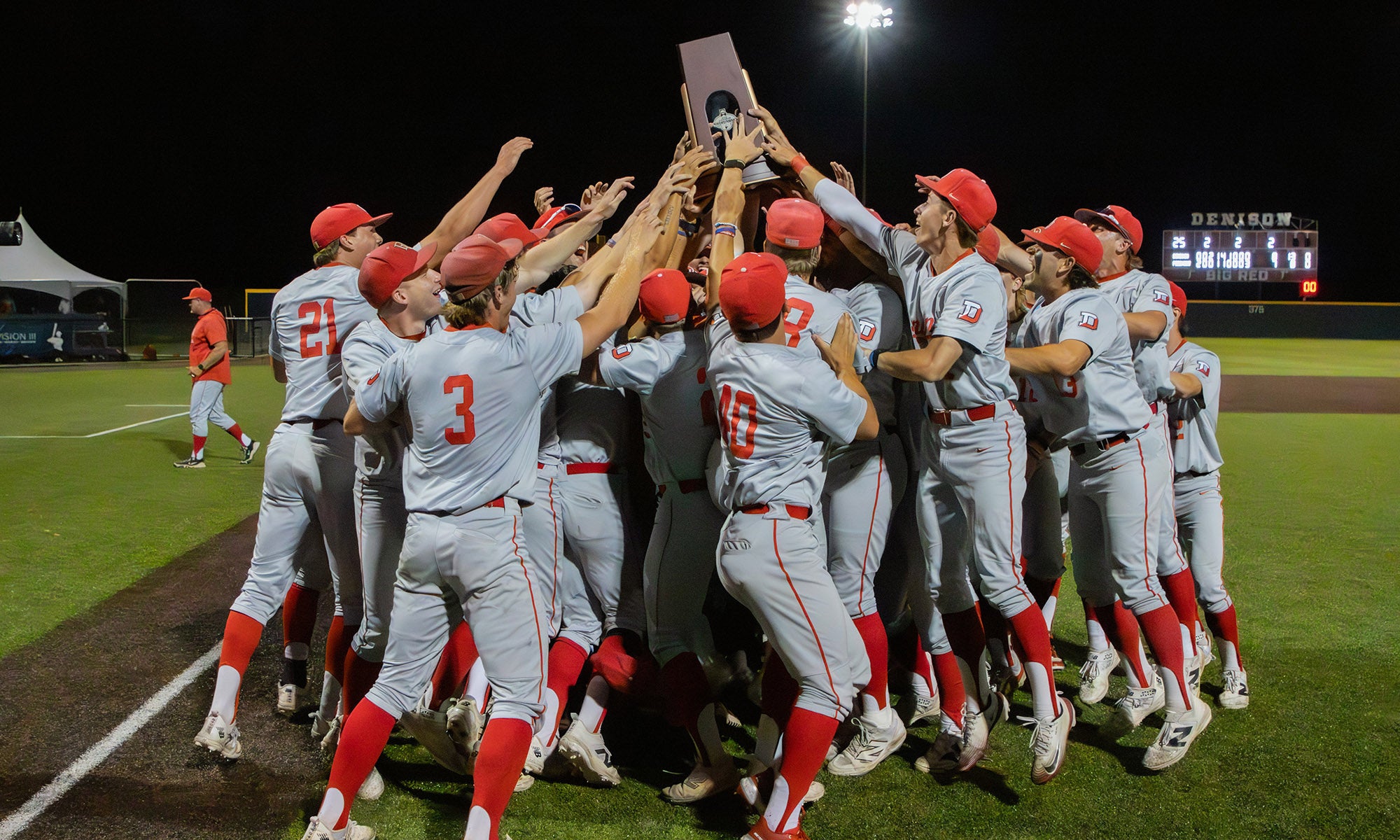 A team of baseball players grouped together on a baseball field at night hold up a championship trophy in celebration.