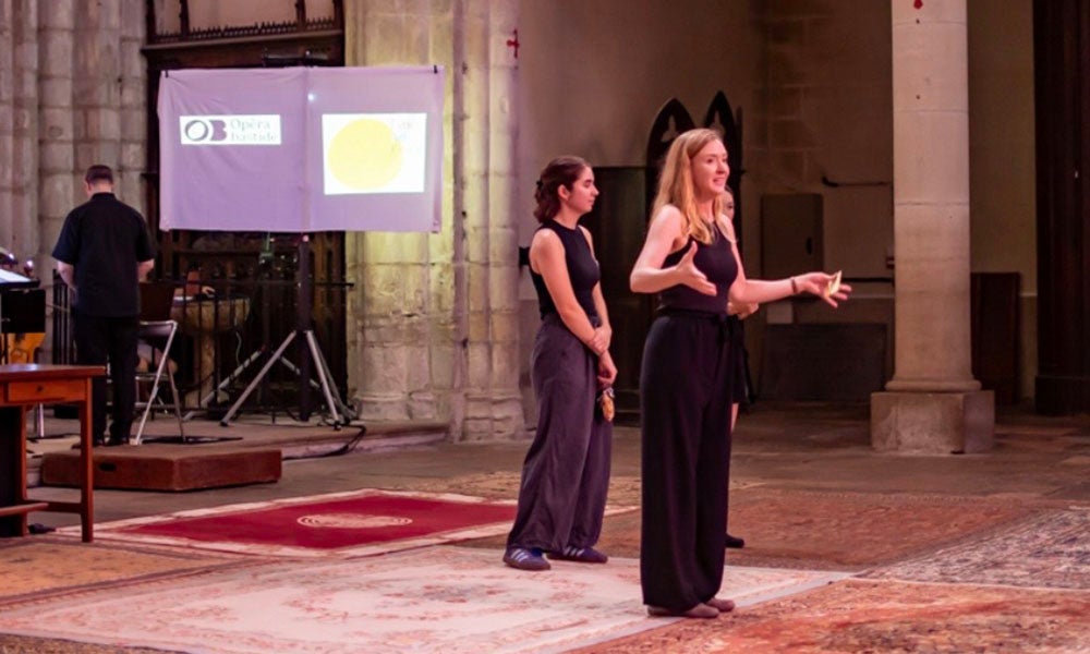 Two women in black outfits stand and gesture while presenting in a large indoor space, with a screen and projector visible in the background.