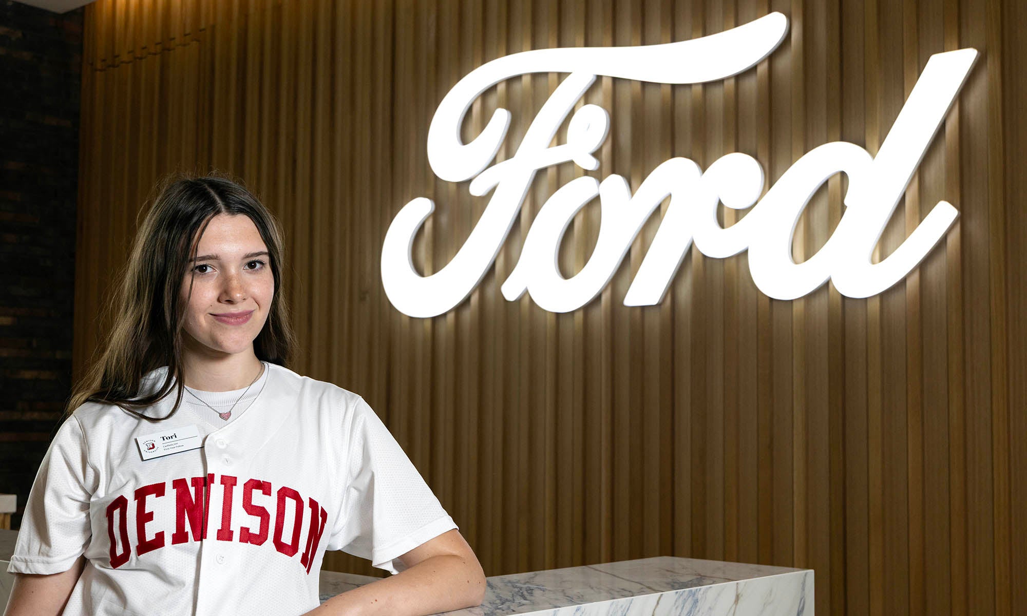 A person in a Denison t-shirt stands in front of a large illuminated Ford logo on a wood-paneled wall.
