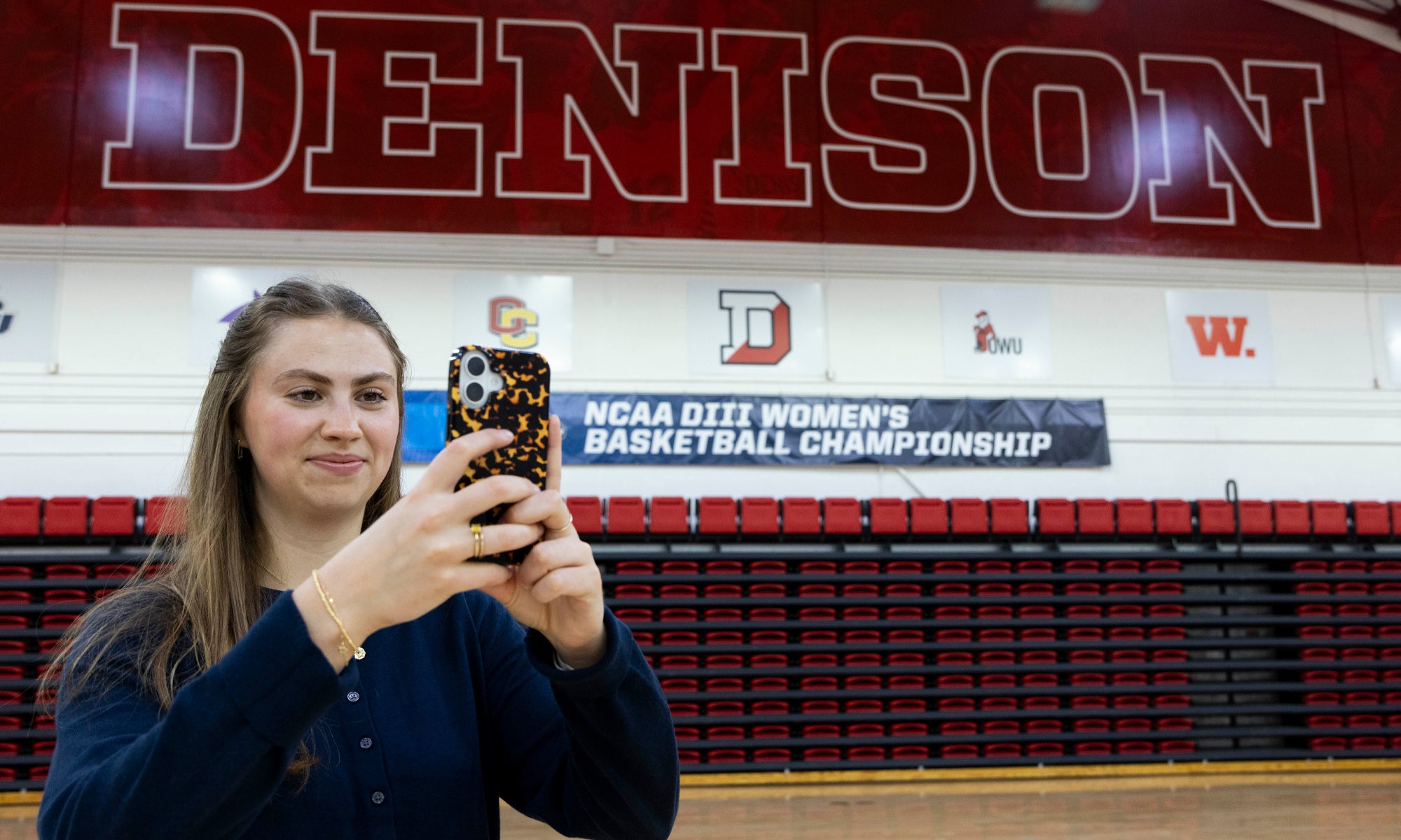 A person uses a phone to record a video inside a gymnasium with DENISON in large letters on the wall and a banner for the NCAA DIII Women's Basketball Championship in the background.