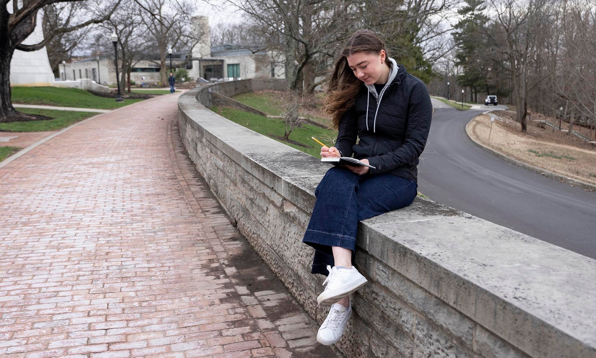 A young woman sits on a curved stone wall along a brick path writing in her notebook.
