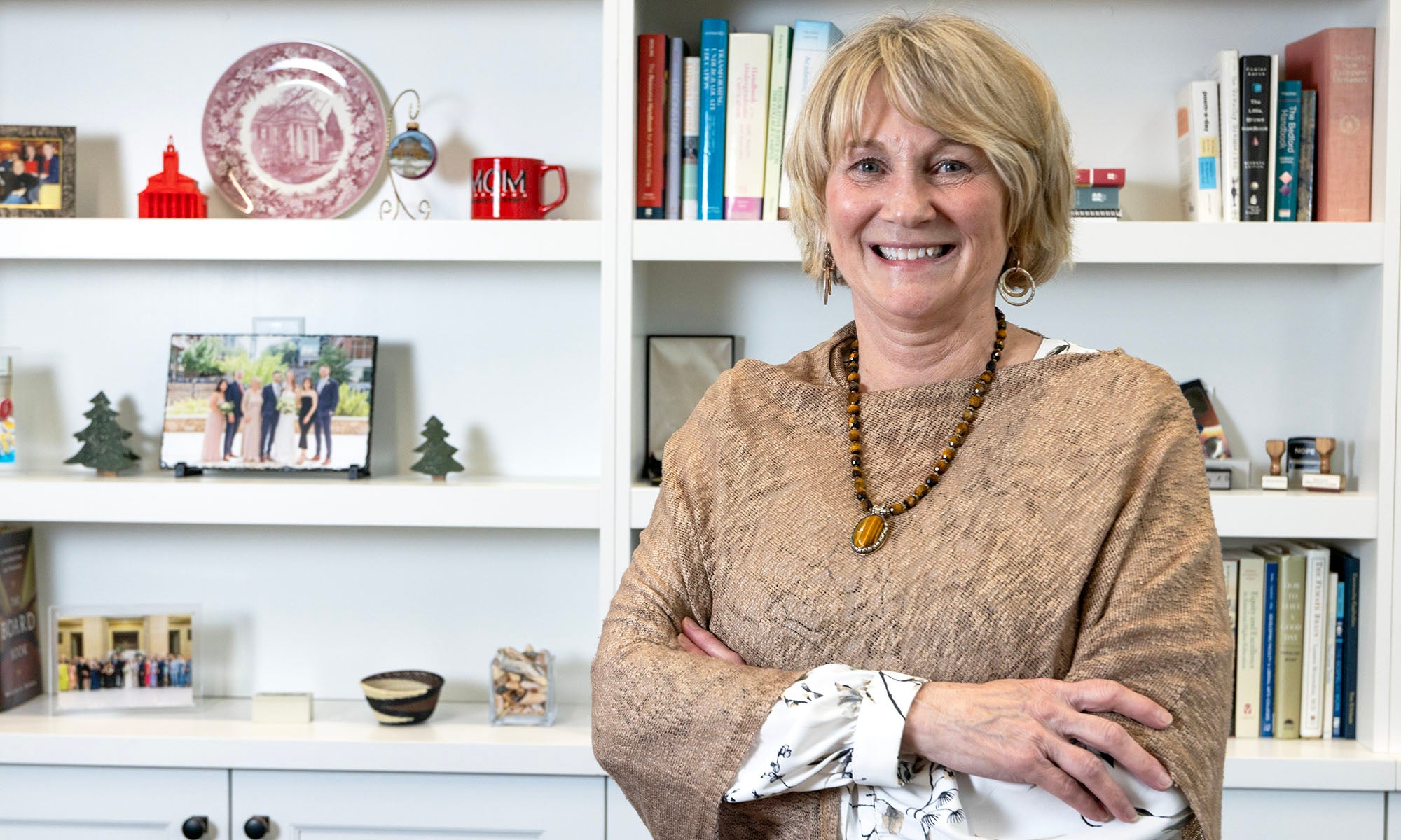 A smiling woman with short blonde hair stands in front of white bookshelves filled with books and decorative items, wearing a textured brown top, a necklace, and earrings, with her arms crossed.