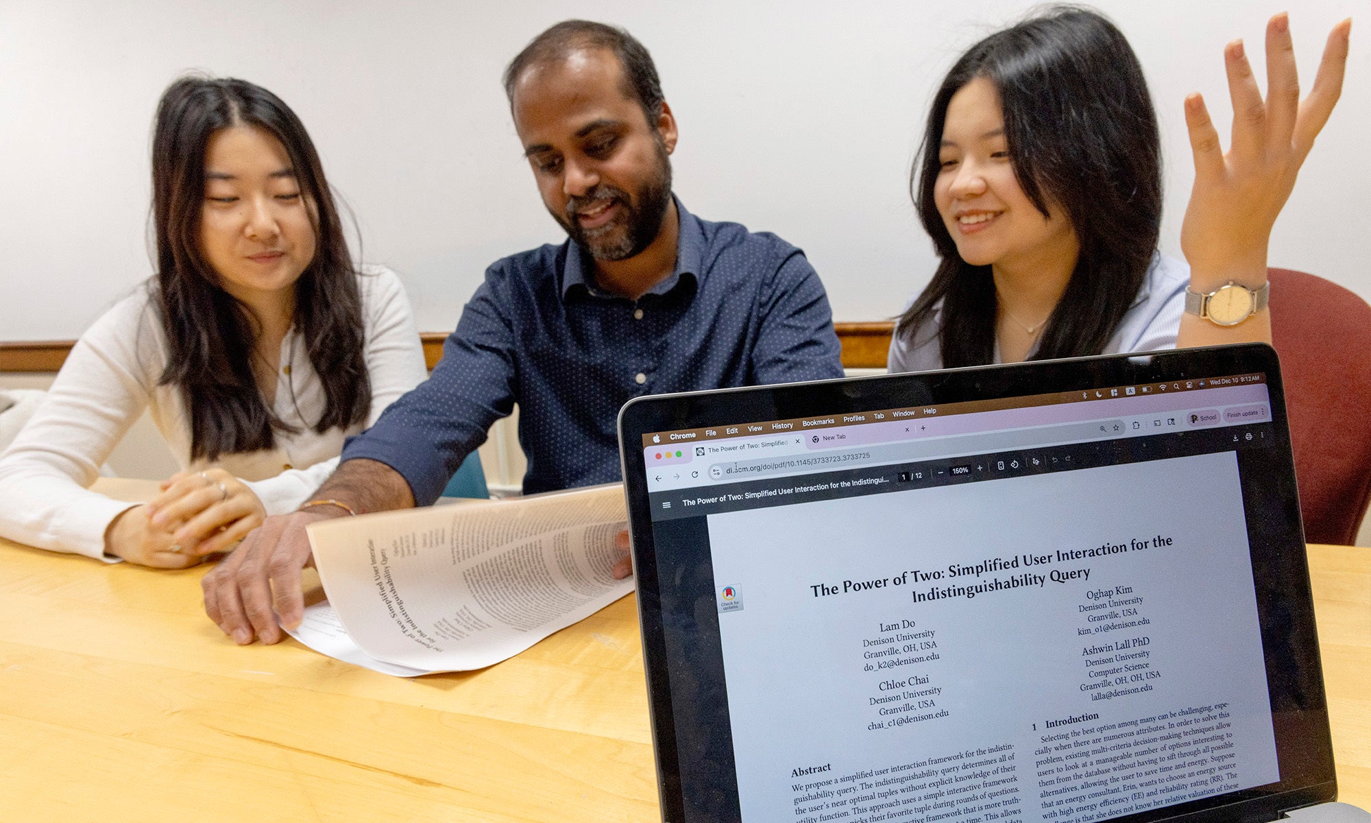 Two college students and a faculty member sit at a table discussing a printed document, which is digitally presented on a laptop screen in the foreground.