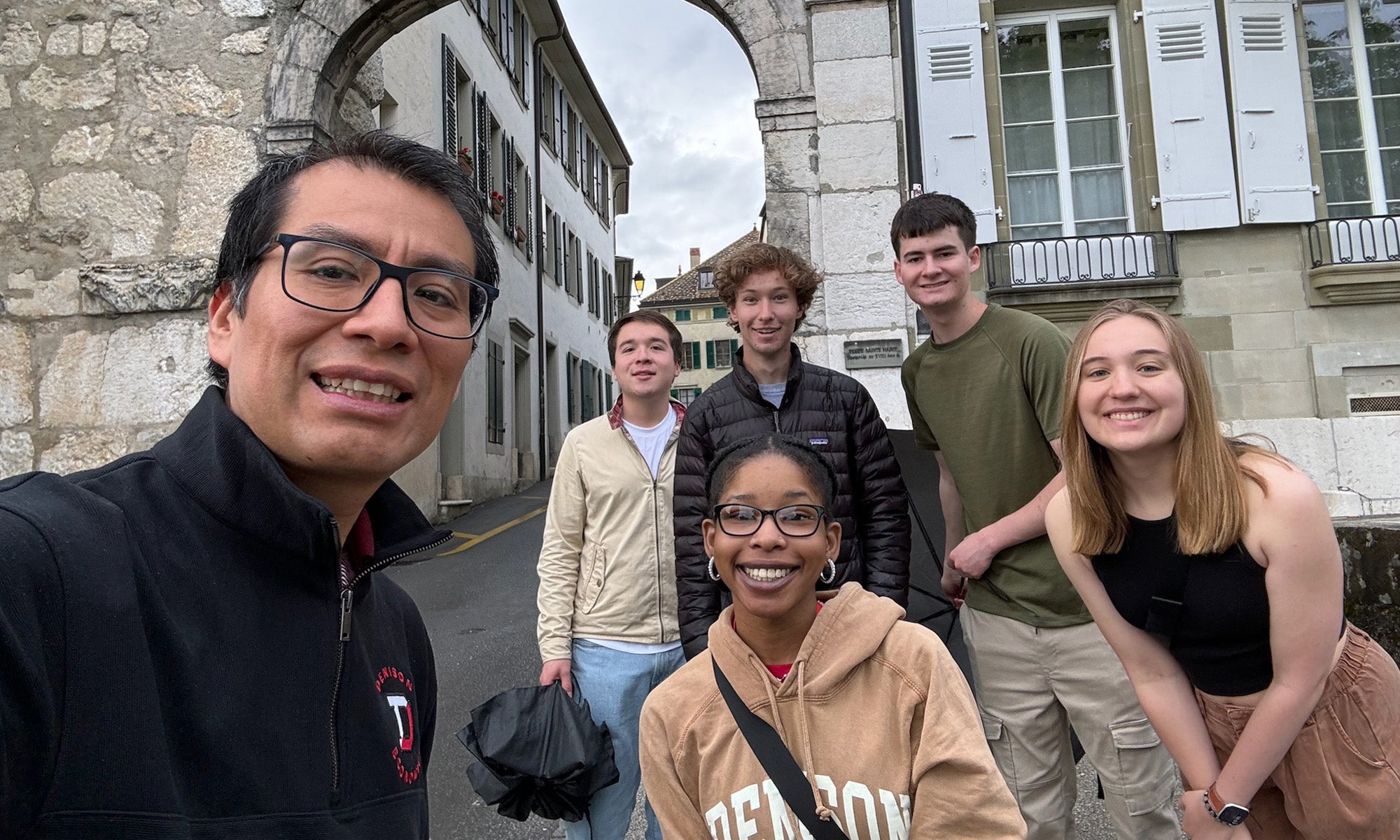 Six people pose and smile for a group photo on a street under a stone archway, with historic buildings in the background.