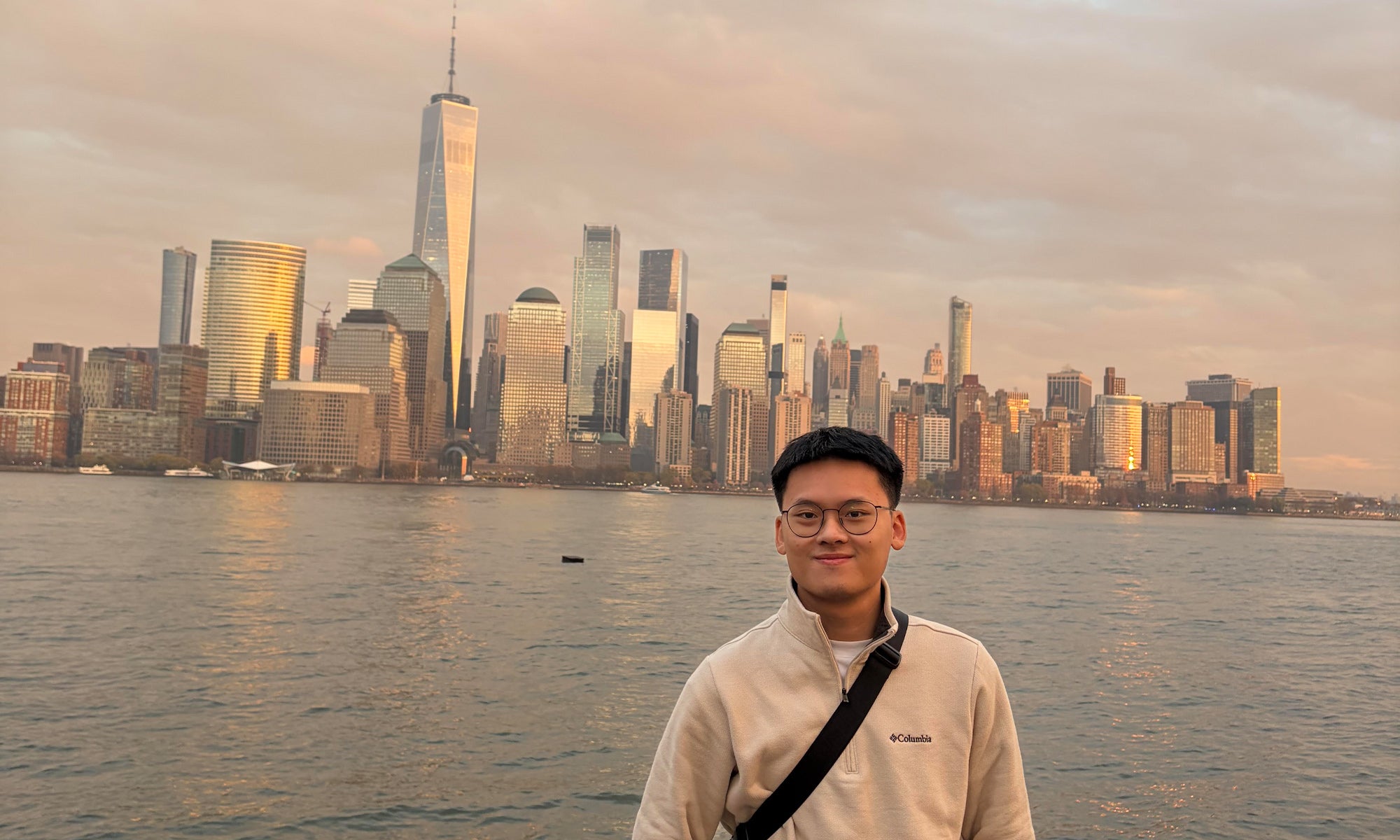 A man wearing glasses and a light jacket stands in front of the New York City skyline by the water at sunset.