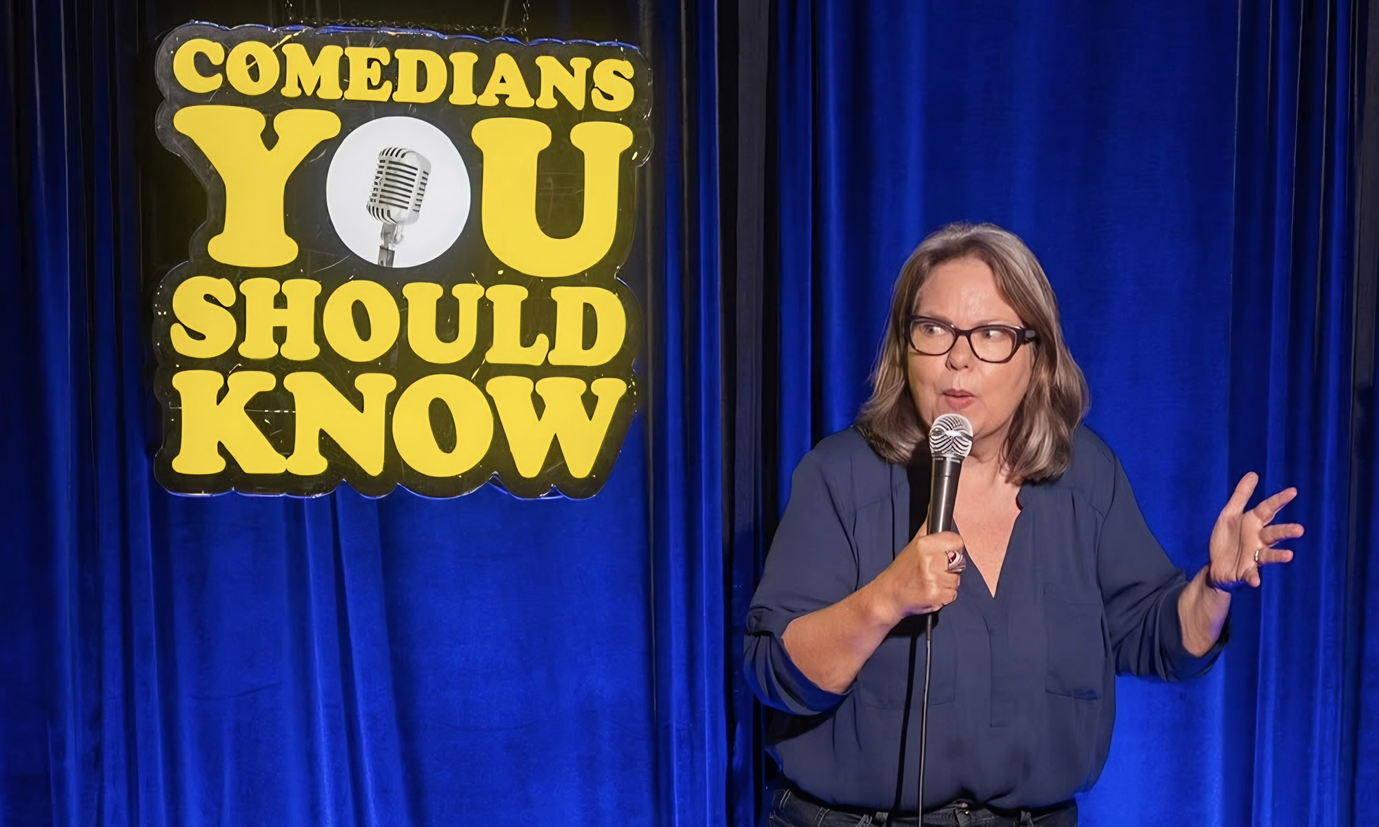 A woman in glasses holds a microphone and gestures while performing in front of a blue curtain with a Comedians You Should Know sign.